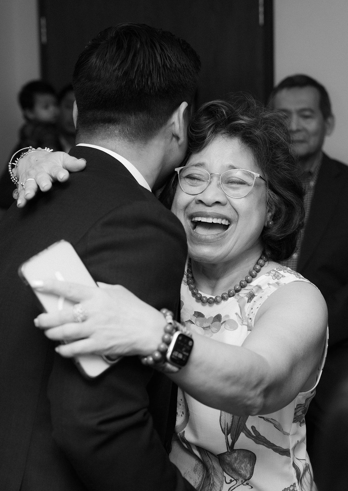 Elderly Asian woman laughing joyfully while embracing a man in a suit at a wedding celebration.
