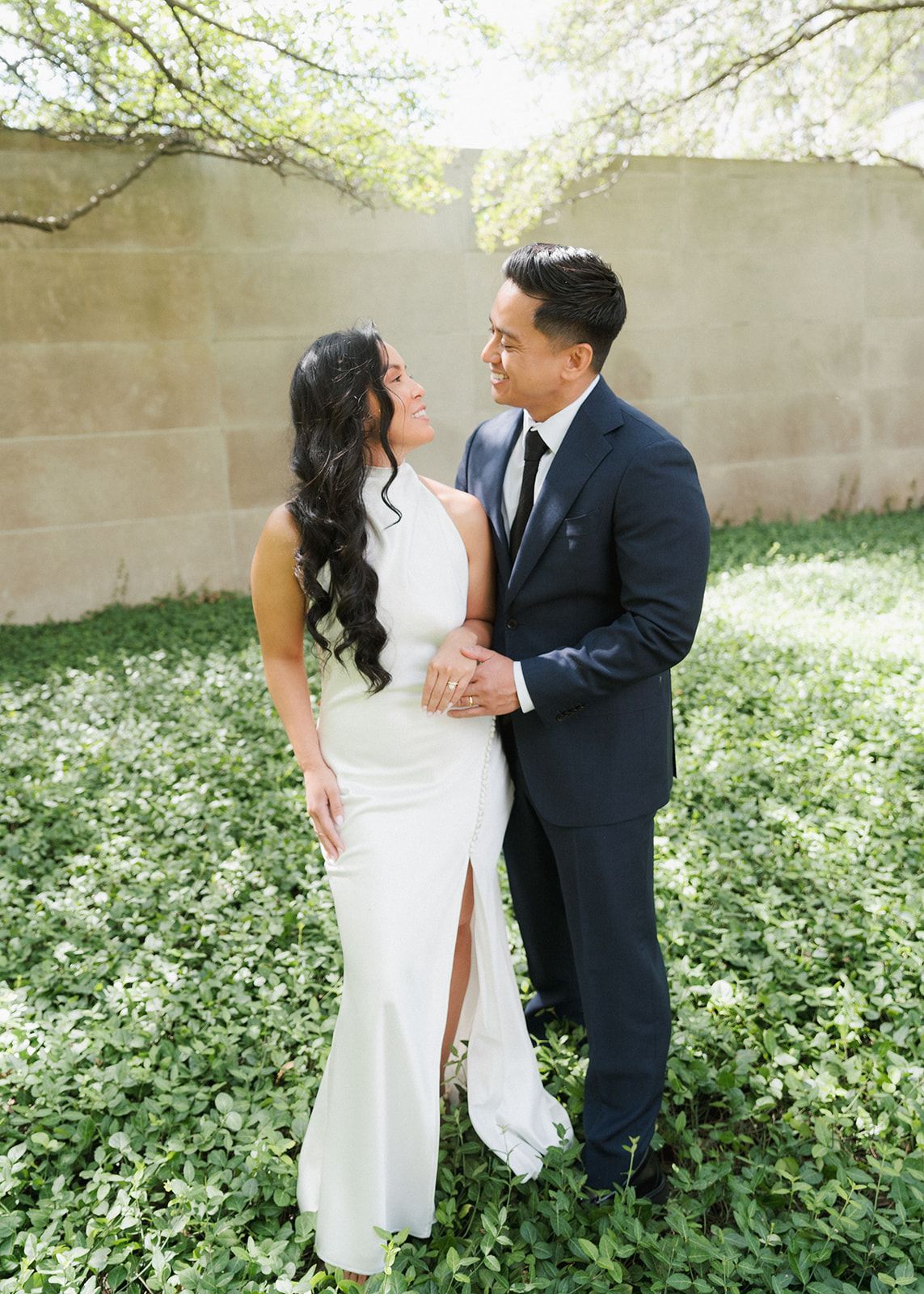 Couple in wedding attire embrace outdoors, woman in white gown and man in navy suit, surrounded by lush green ground cover.