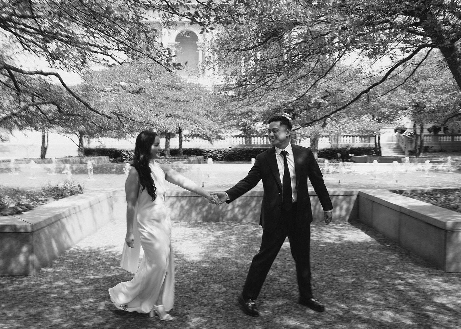Black and white photo of bride and groom holding hands walking under blooming cherry blossom trees.