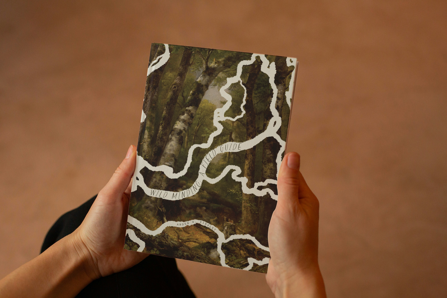 Hands hold a book with a painting of a forest and a winding river showcasing a story of re-wilding, inclusivity, and decolonization.