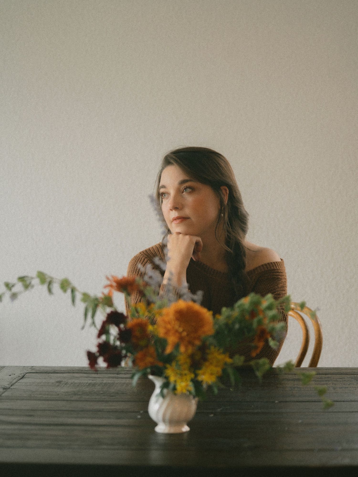 A woman sits with her chin on her hand behind a pitcher of autumn wildflowers taken during a fall portrait session.
