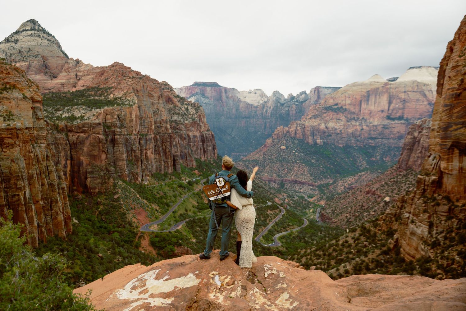Couple standing on rocky overlook admiring stunning Zion Canyon valley views with red cliffs and green trees below.