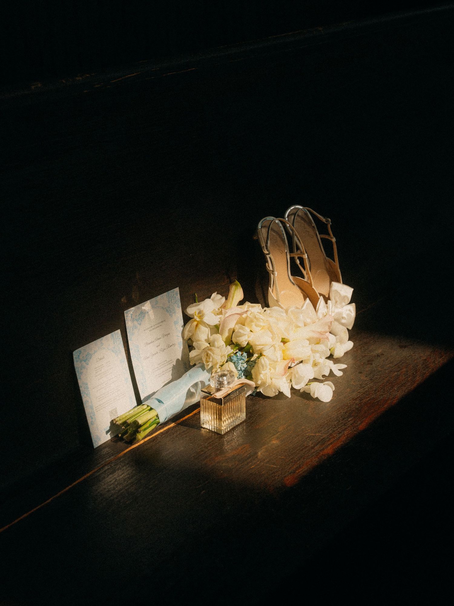 Elegant wedding flat lay with white floral bouquet, clear heels, and stationery on a dark wooden surface.