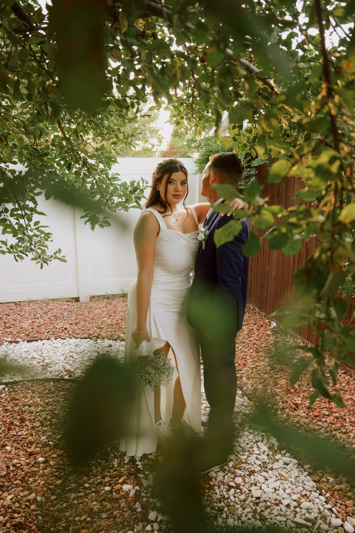 A bride and groom share an intimate moment framed by lush green leaves in a garden setting.