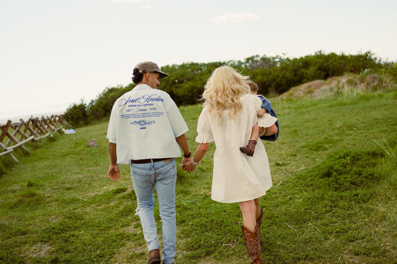 Couple walking hand-in-hand on green hillside, man in white tee and jeans, woman in white dress and brown boots.