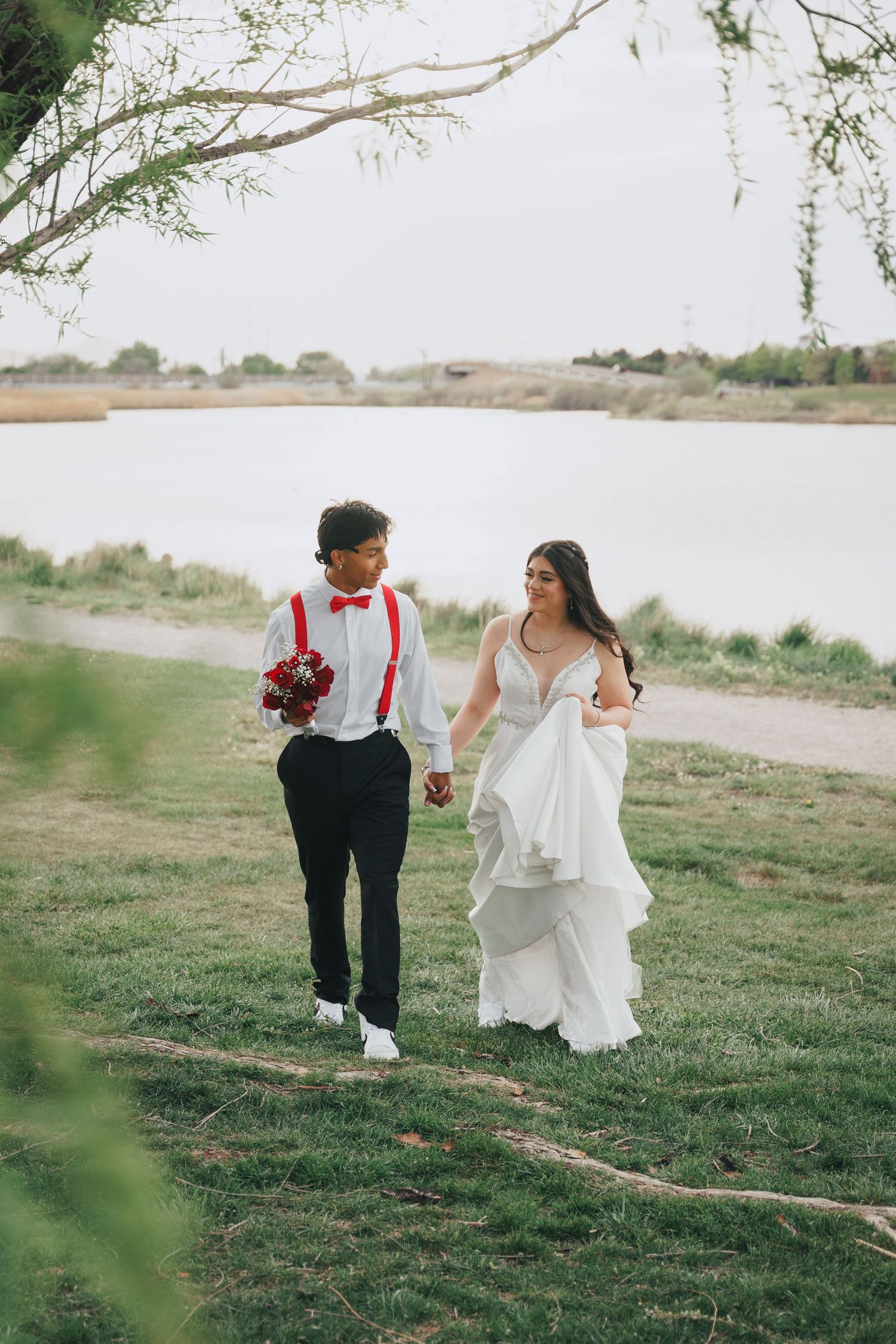A bride and groom walk hand in hand along a grassy lakeside path, smiling at each other.
