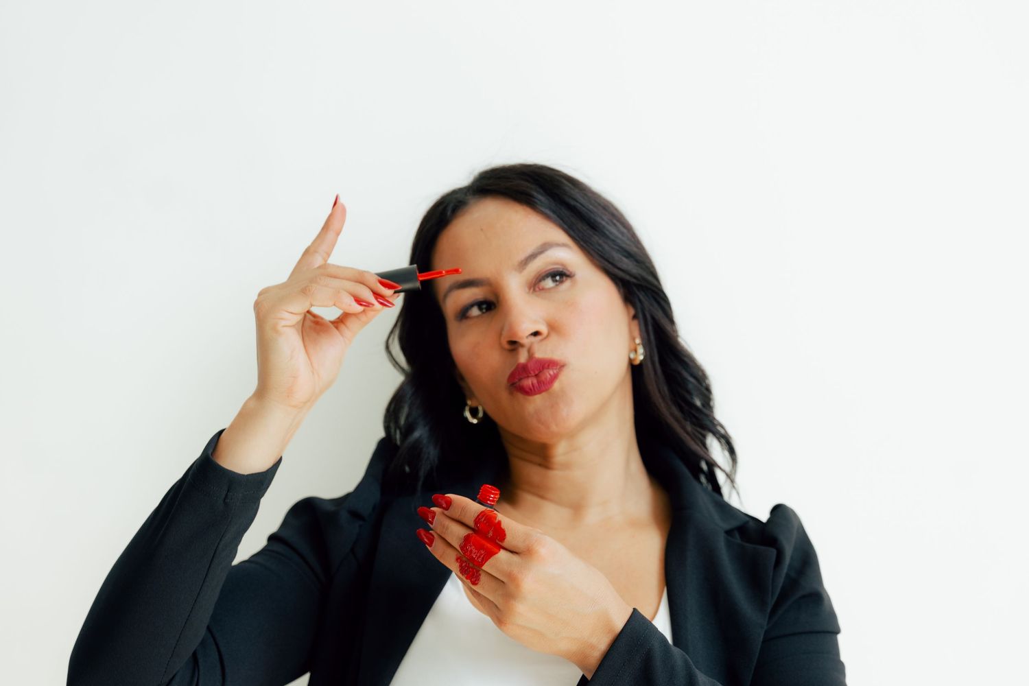 Confident woman in black blazer playfully applying red lipstick against white background.