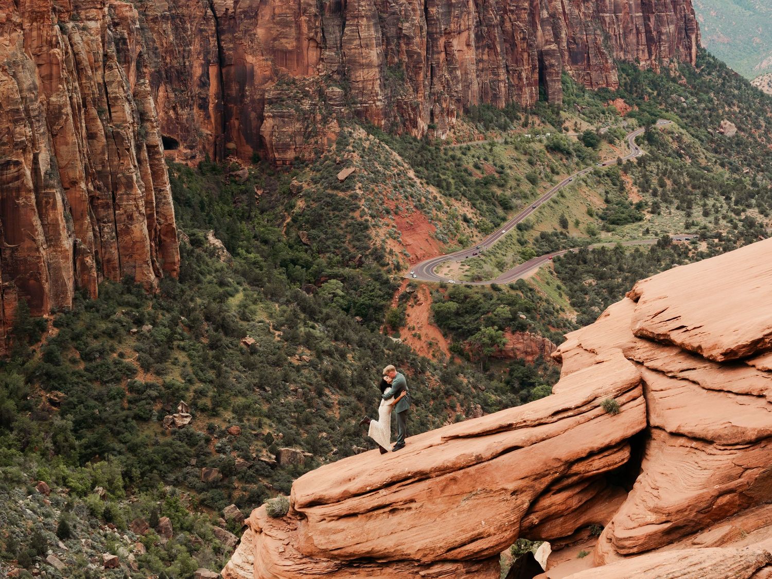 Hiker stands on red sandstone cliff edge overlooking vast Zion National Park canyon with layered rock walls and winding road.