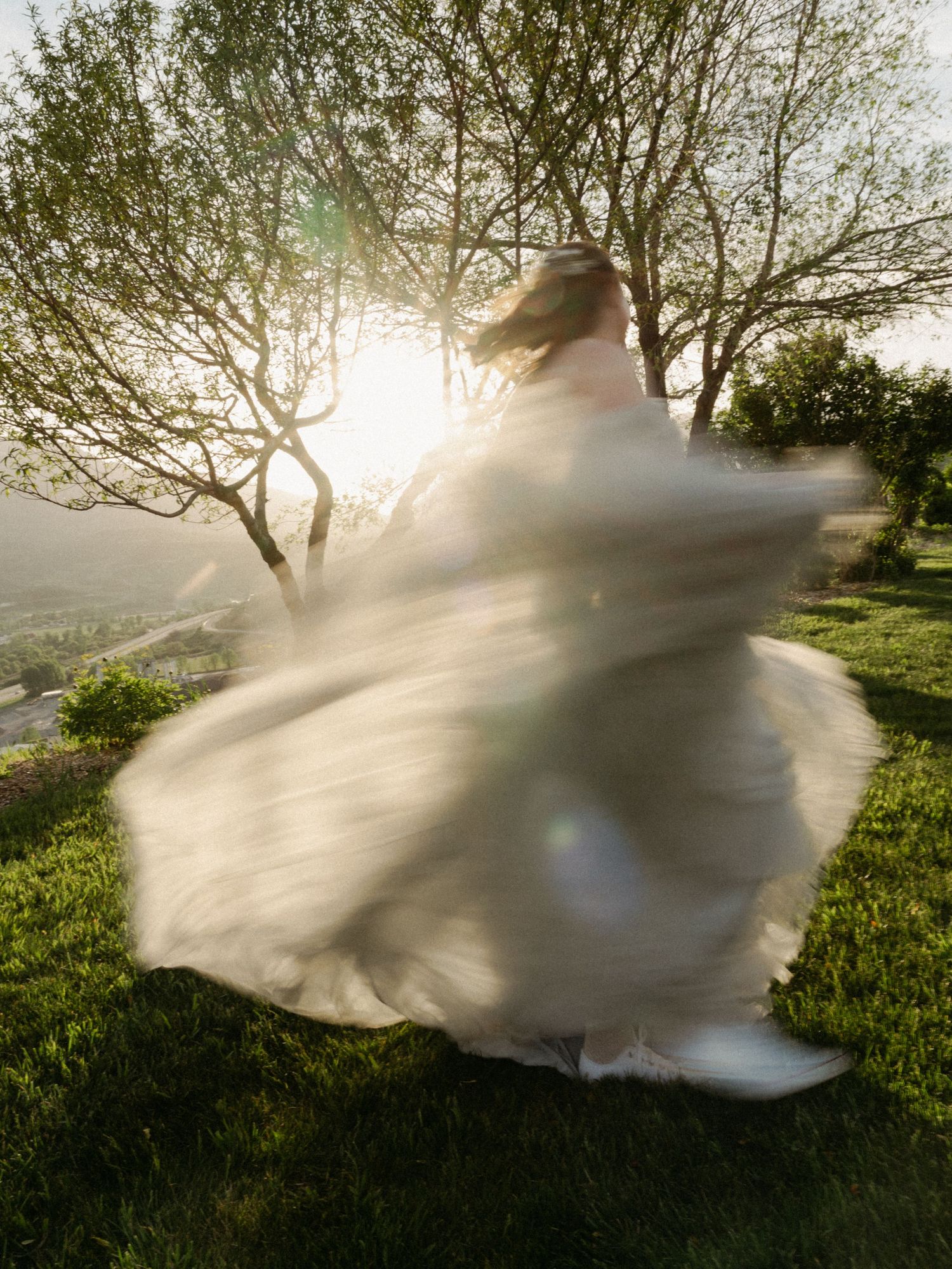 Bride in white gown twirling outdoors at golden hour, motion blur capturing flowing dress movement against sunlit trees.