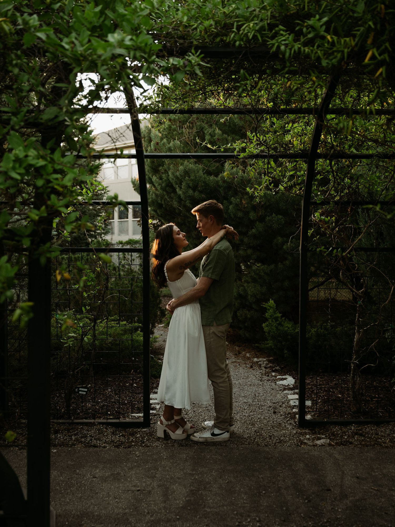 Couple embracing under a garden arch surrounded by lush green foliage, woman in white dress, romantic outdoor setting.