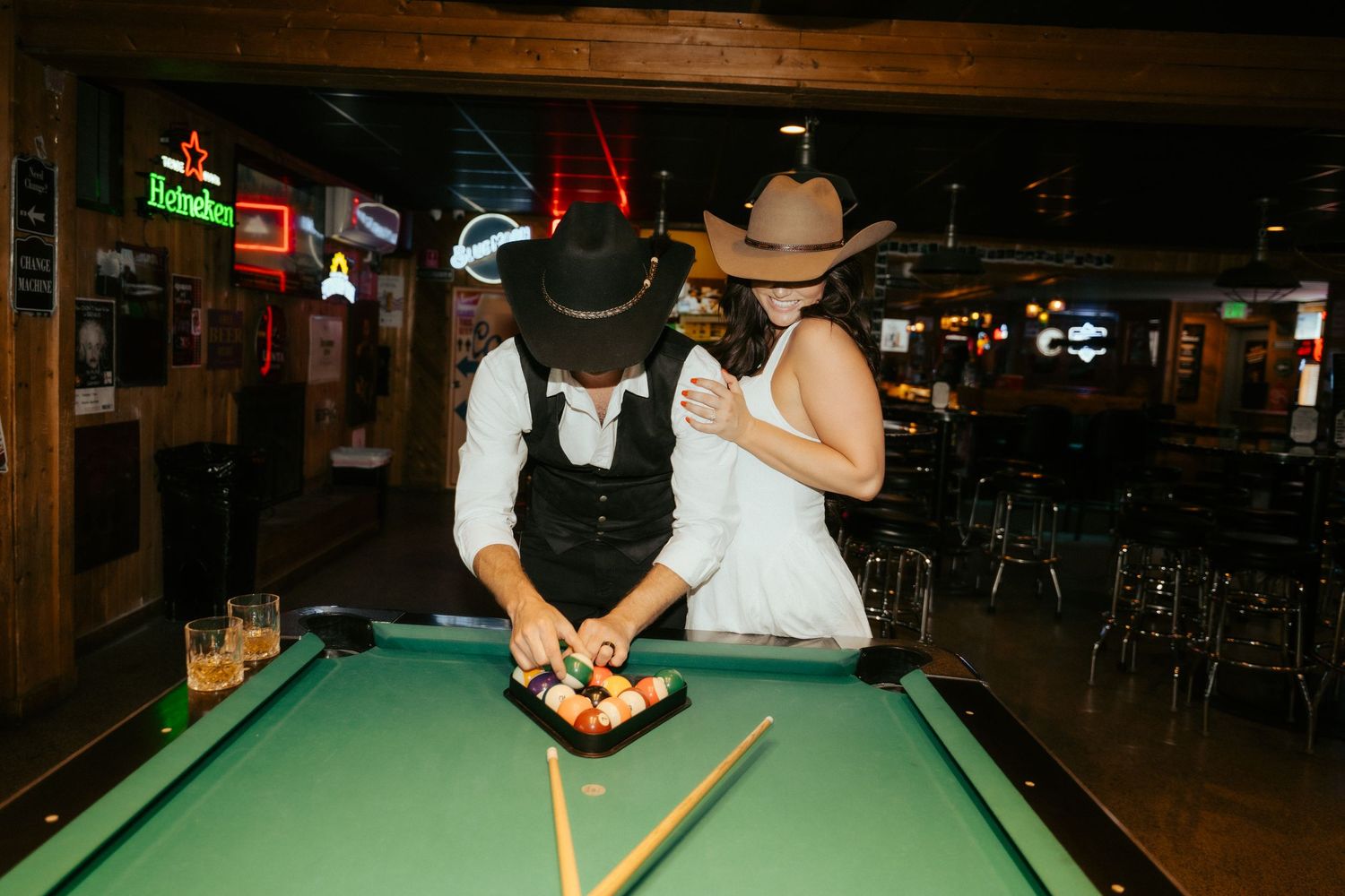 Couple in cowboy hats playing pool at a rustic bar, leaning over a green billiard table together.