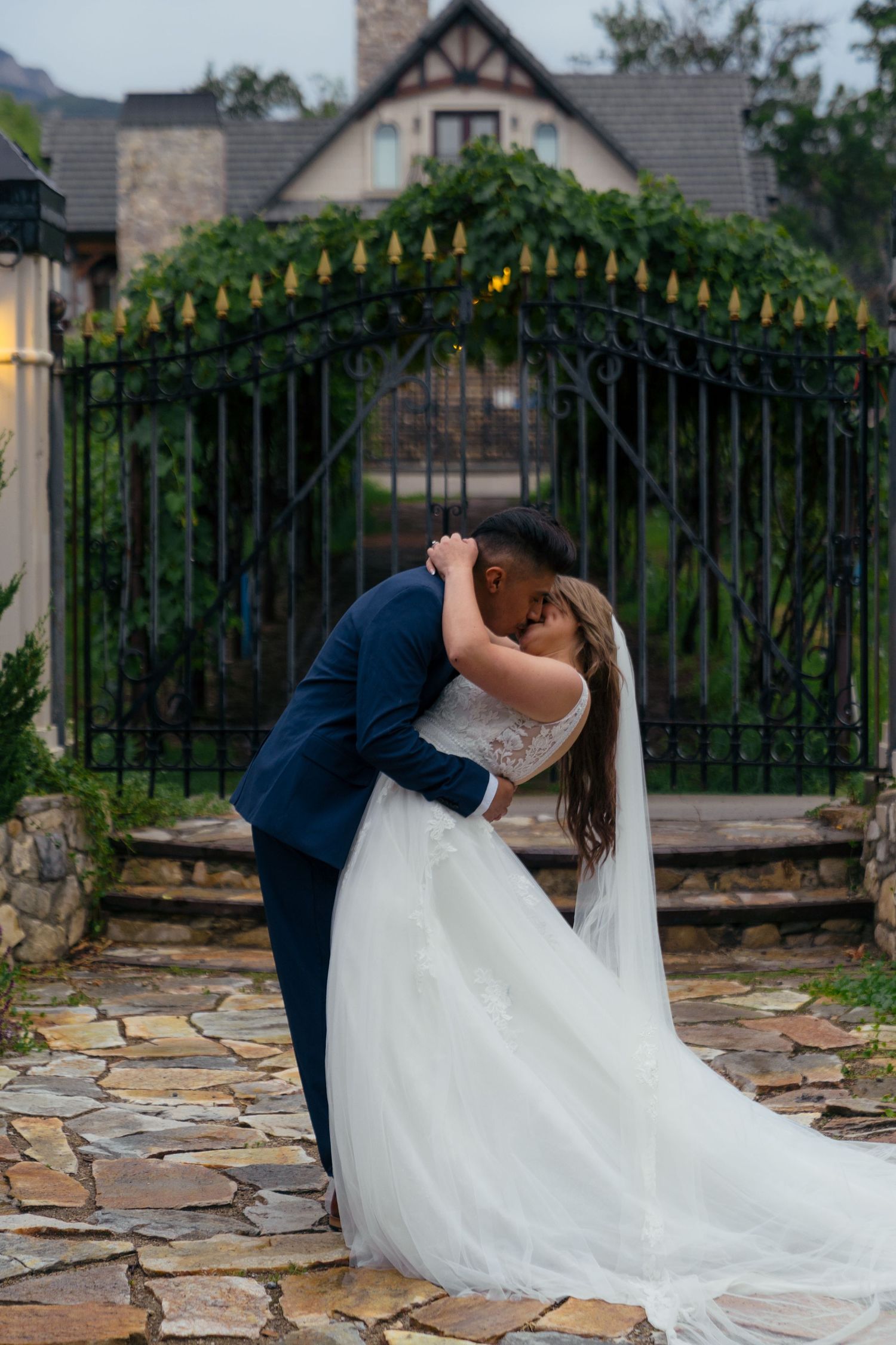 Bride and groom sharing a kiss at dusk before an ivy-covered iron gate at a wedding venue.