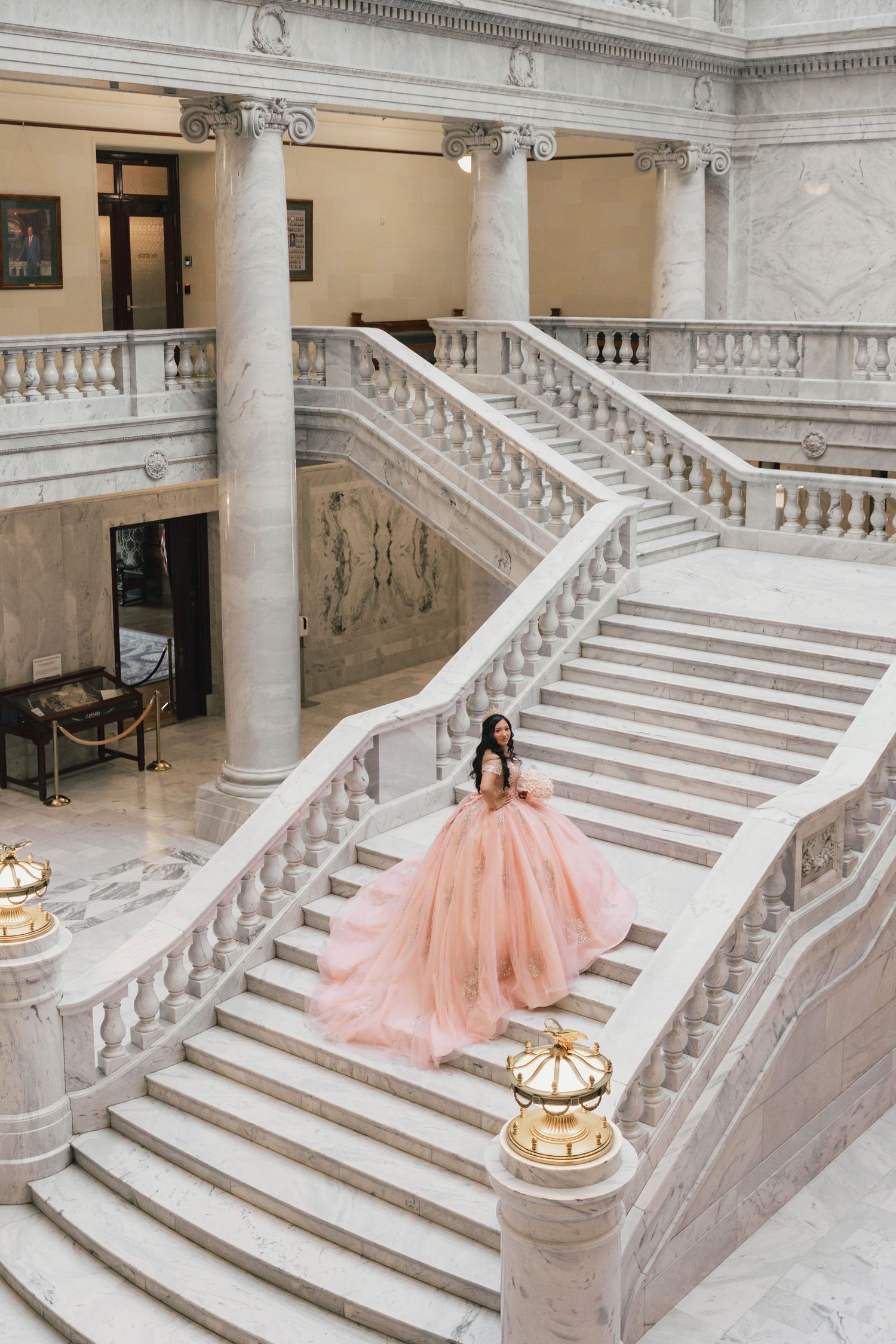 Young woman in a pink tulle ball gown descending a grand marble staircase in an elegant building.