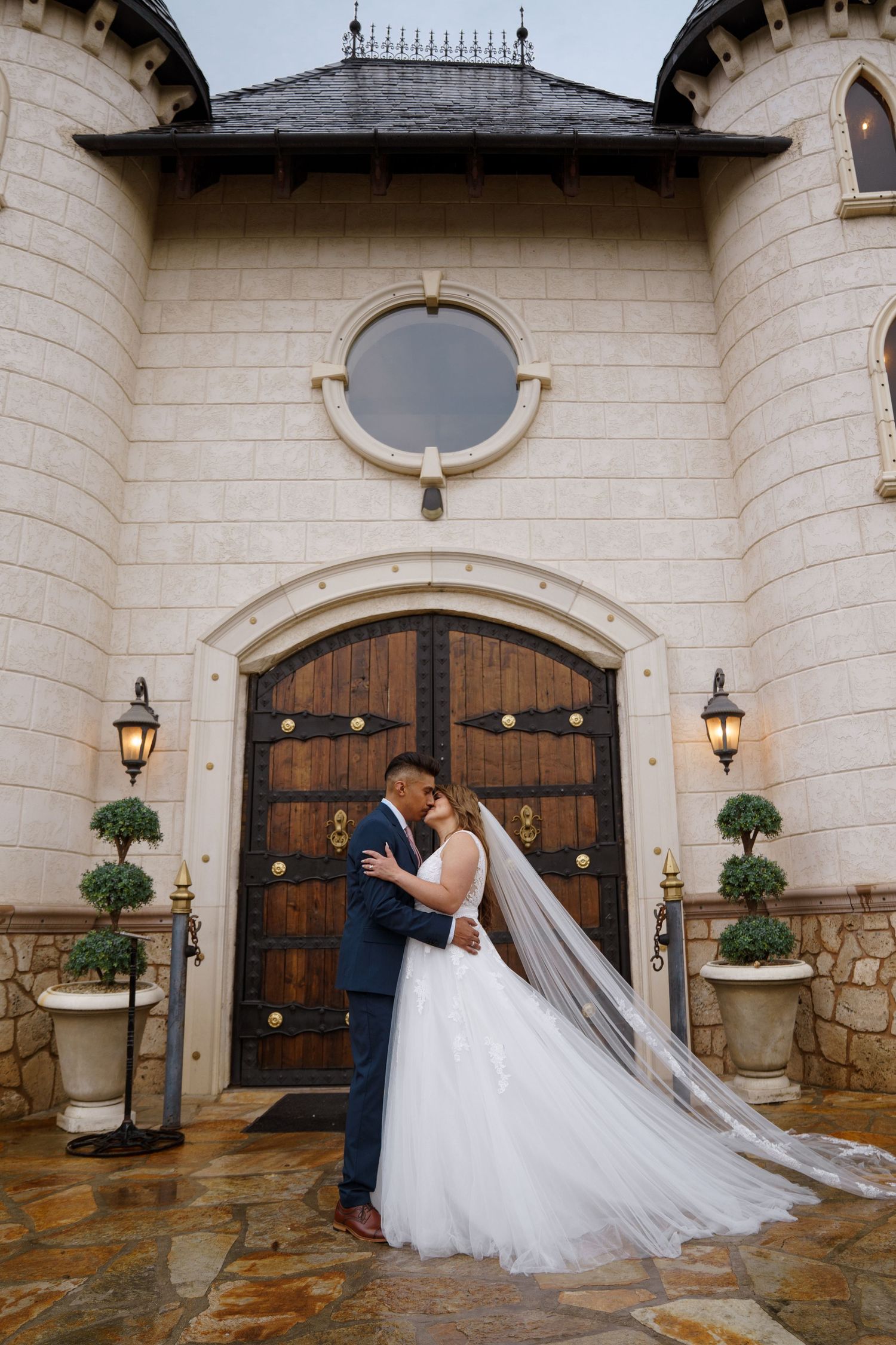 Bride and groom sharing romantic kiss before ornate wooden doors of castle wedding venue.