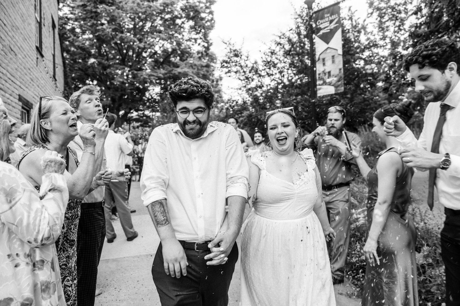 Black and white photo of a joyful couple holding hands walking through cheering wedding guests outdoors.