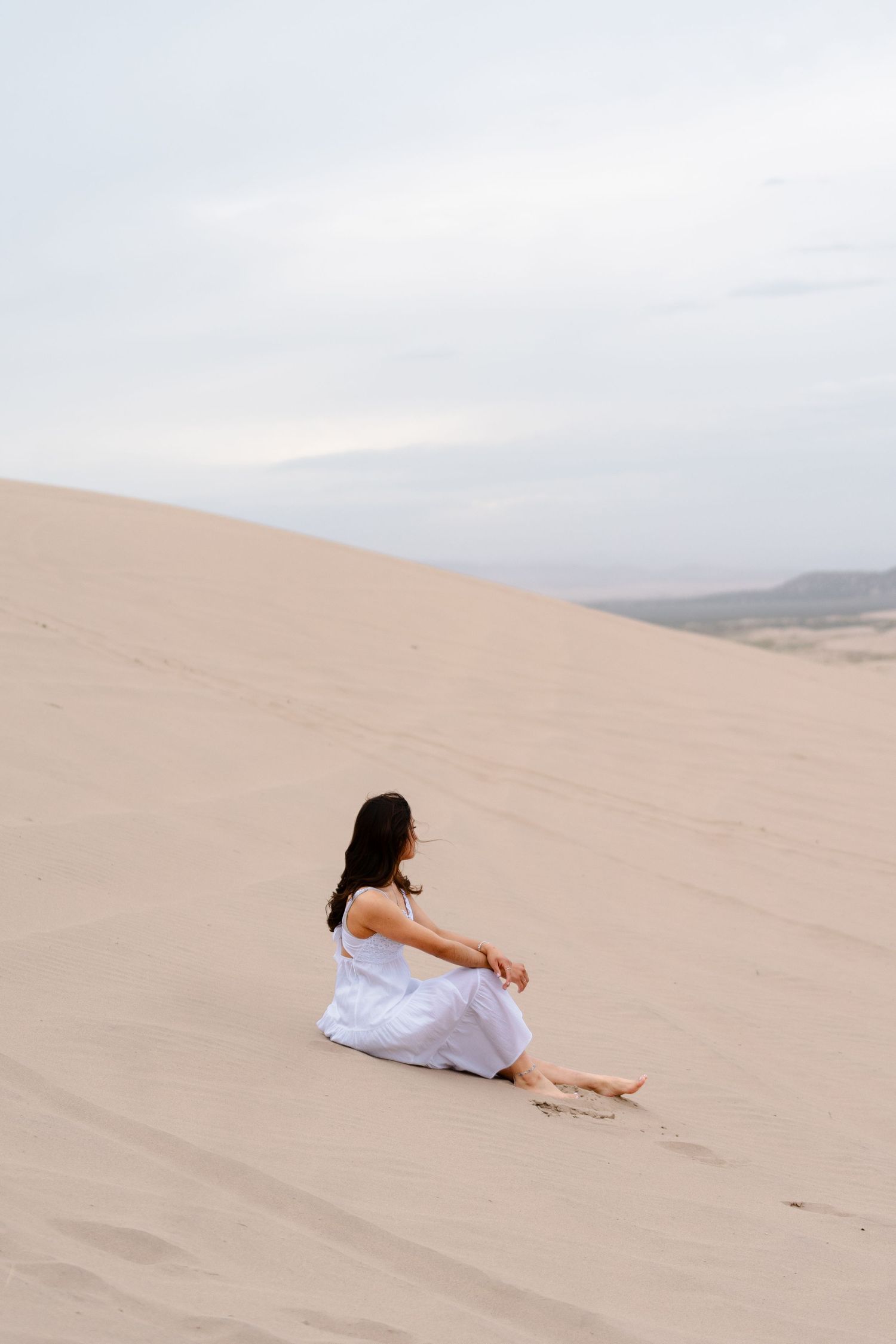 A woman in a white dress sits barefoot on a sandy desert dune under a cloudy sky.