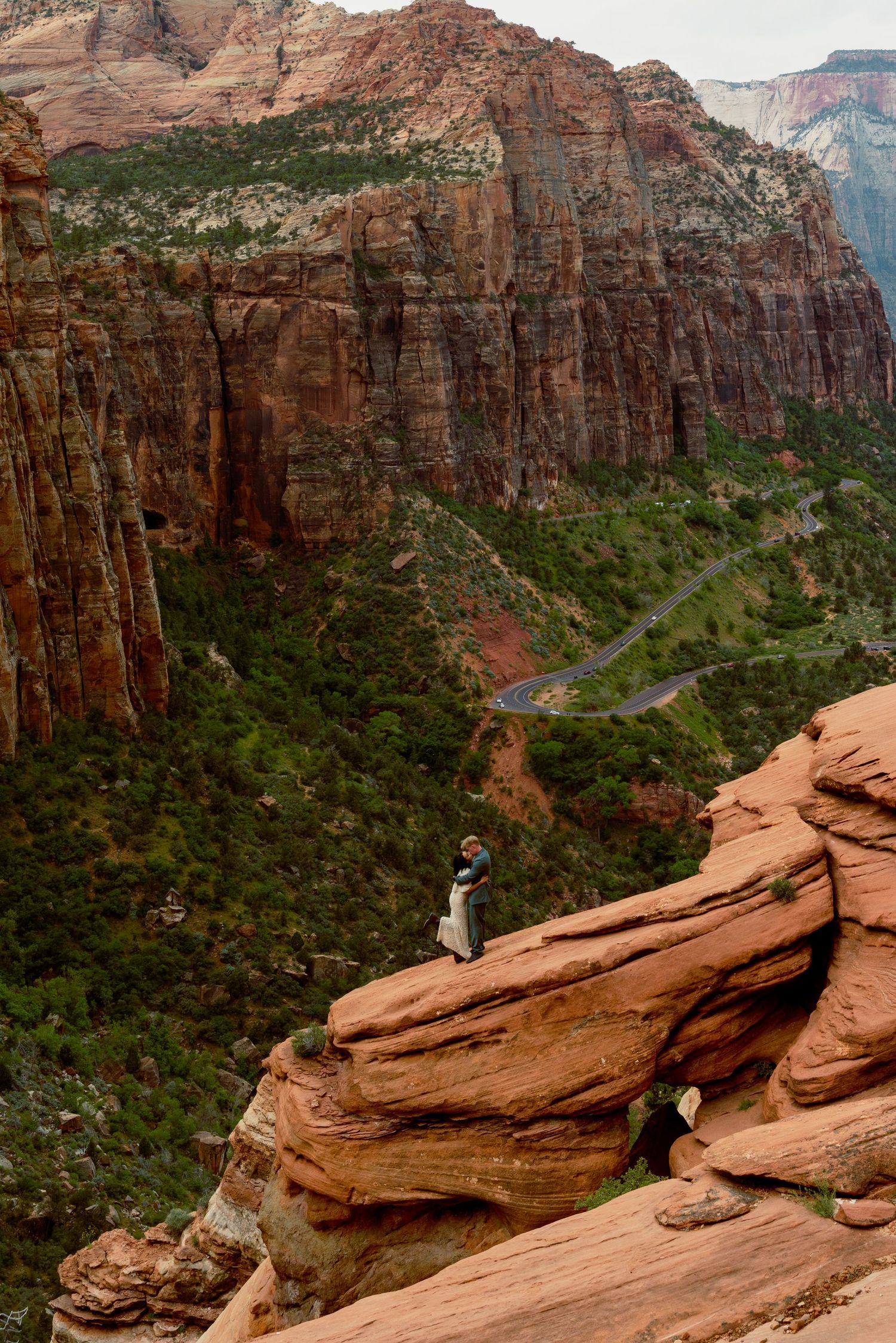 Person standing on red rock ledge overlooking Zion Canyon's dramatic cliffs and winding road below.