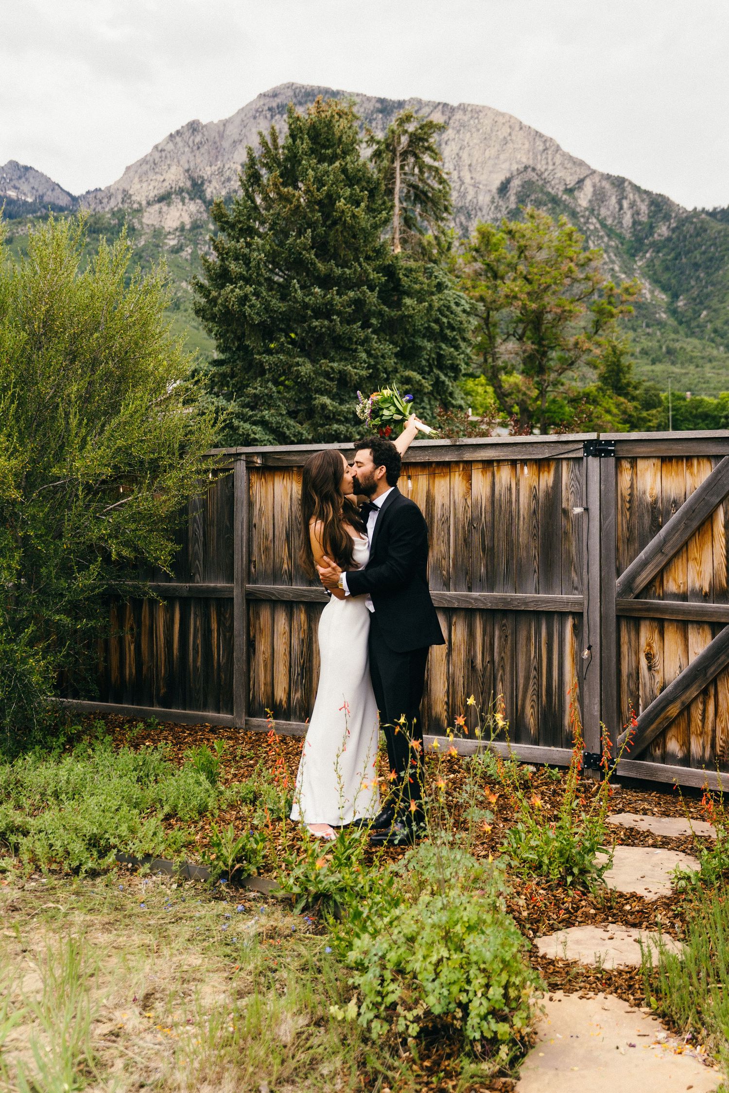Bride and groom kissing in front of rustic wooden fence with mountain backdrop on their wedding day.