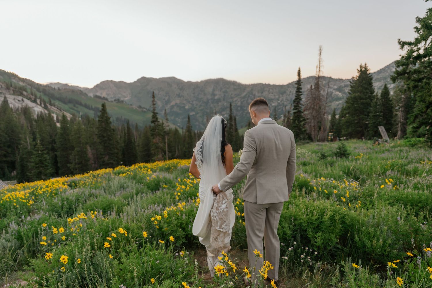 Bride and groom holding hands walking through wildflower meadow with mountain and pine tree backdrop at sunset wedding.