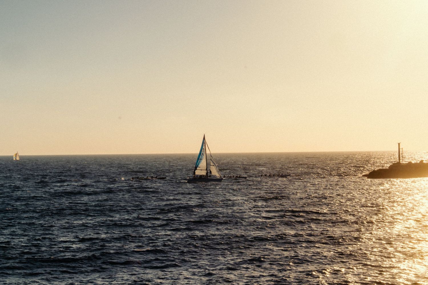 Sailboat sailing on calm ocean waters at golden sunset with another vessel visible on the horizon.