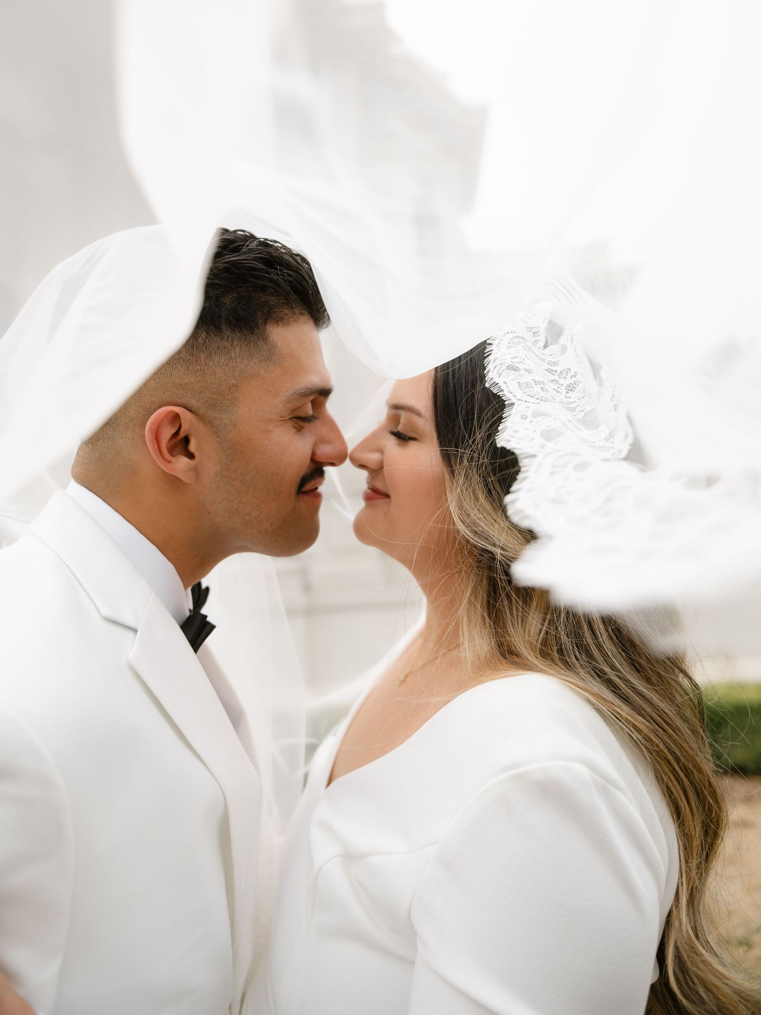 Bride and groom in white attire share a romantic nose-to-nose moment, veil flowing in the wind.