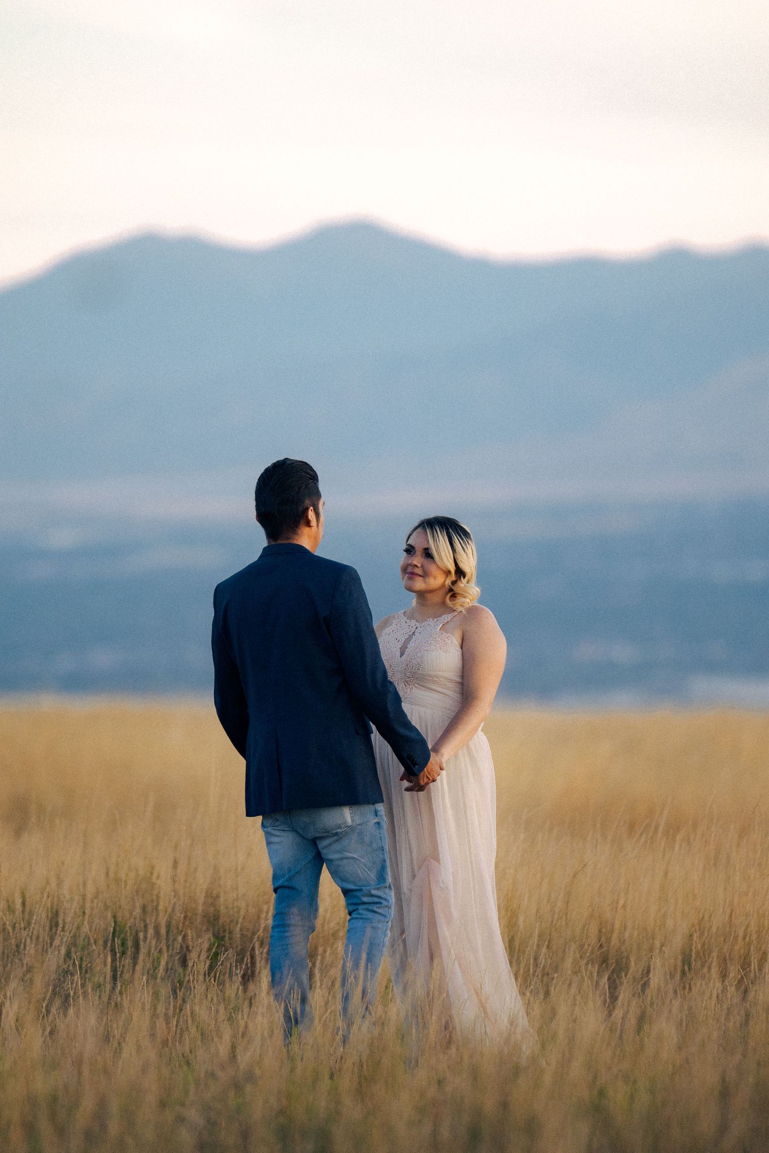 A couple holds hands in a golden field with misty mountains at sunset.