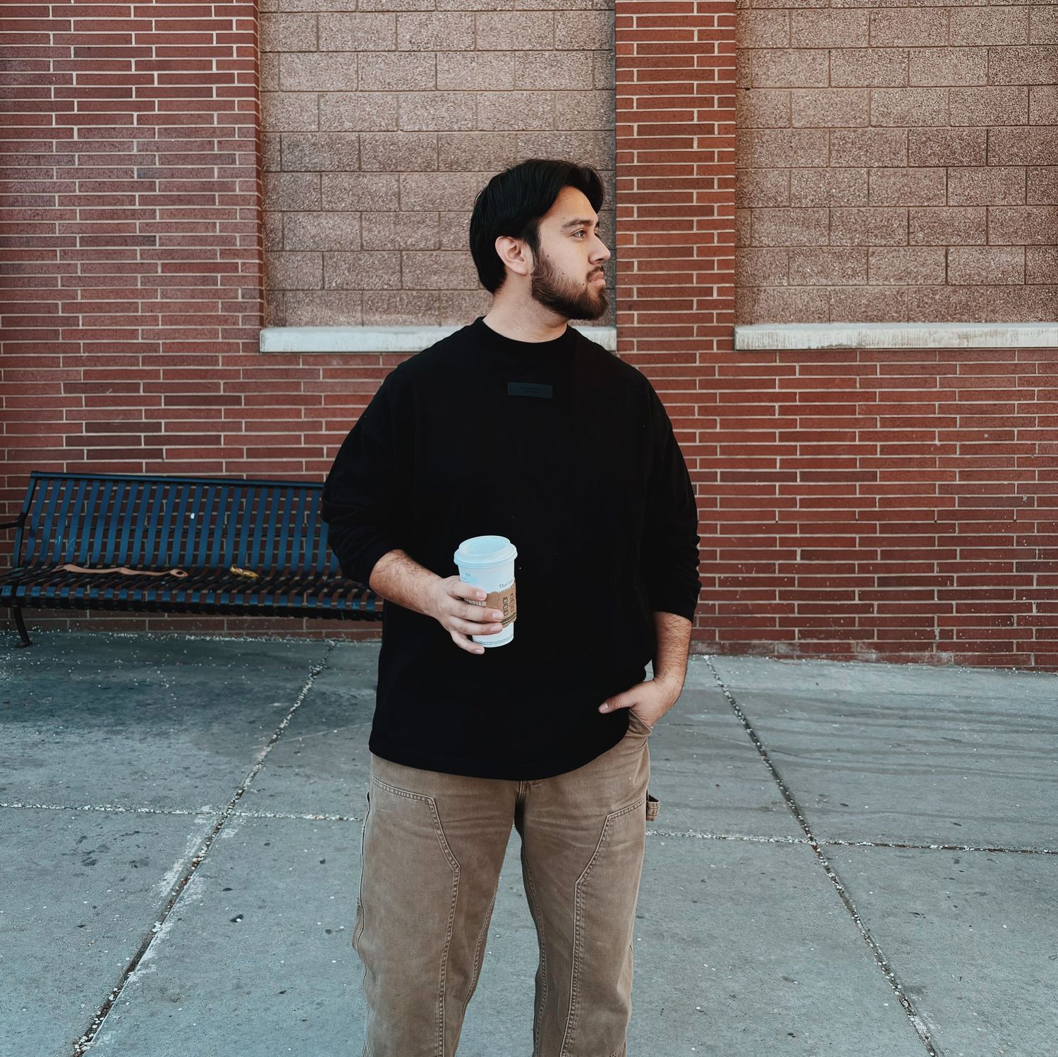 Young man in black oversized shirt holding coffee cup standing near brick wall outdoors.