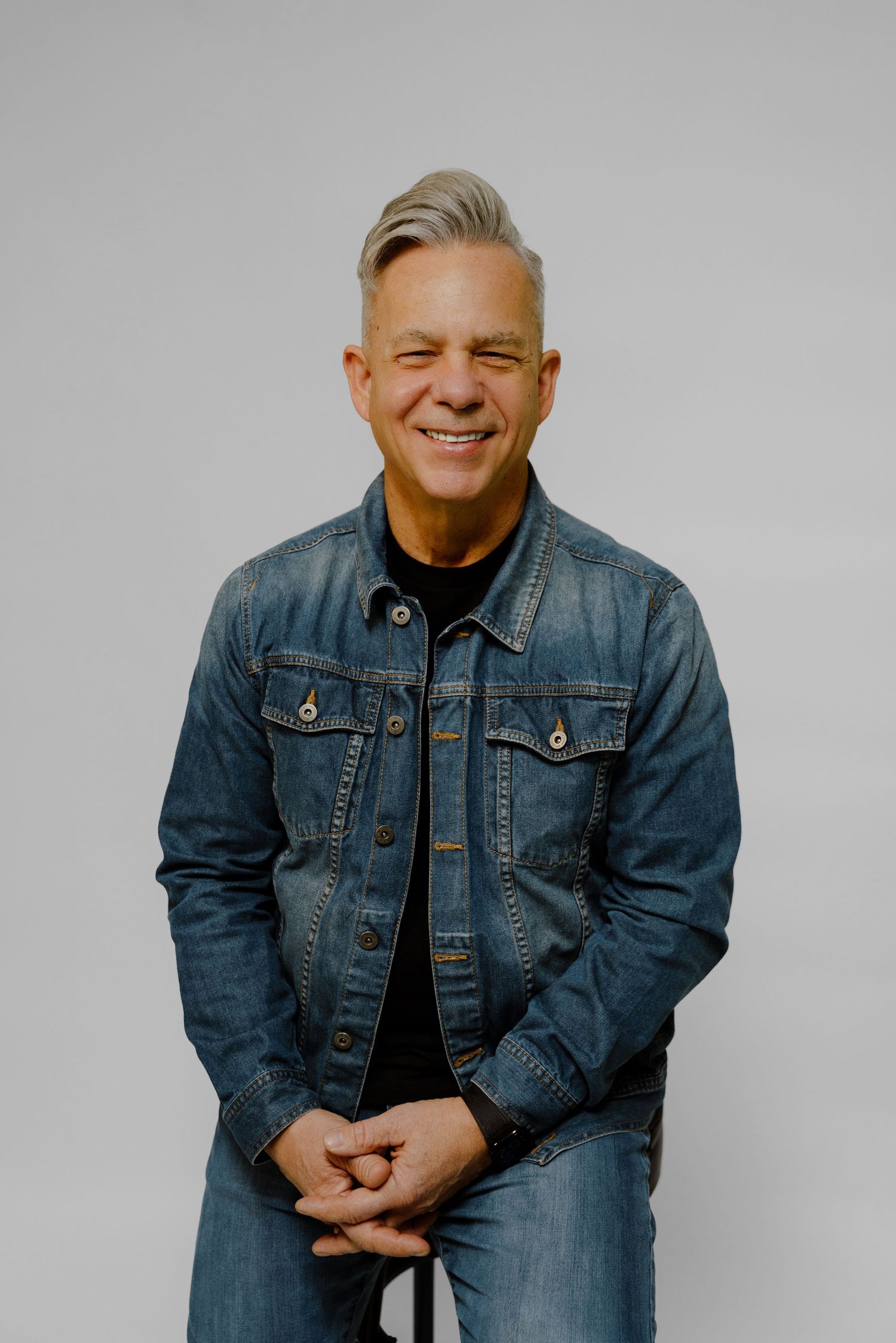 Smiling middle-aged man with gray hair wearing denim jacket seated against light gray background.
