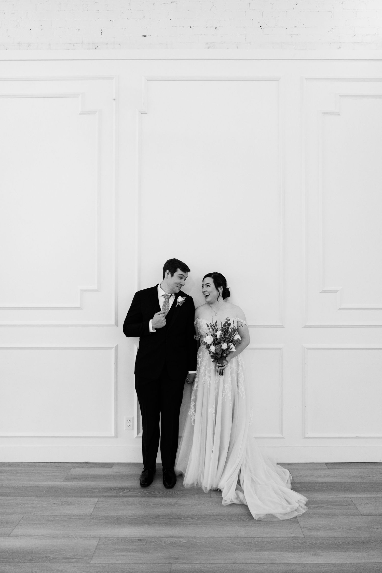 Bride and groom posing together in black and white photo against white paneled wall.