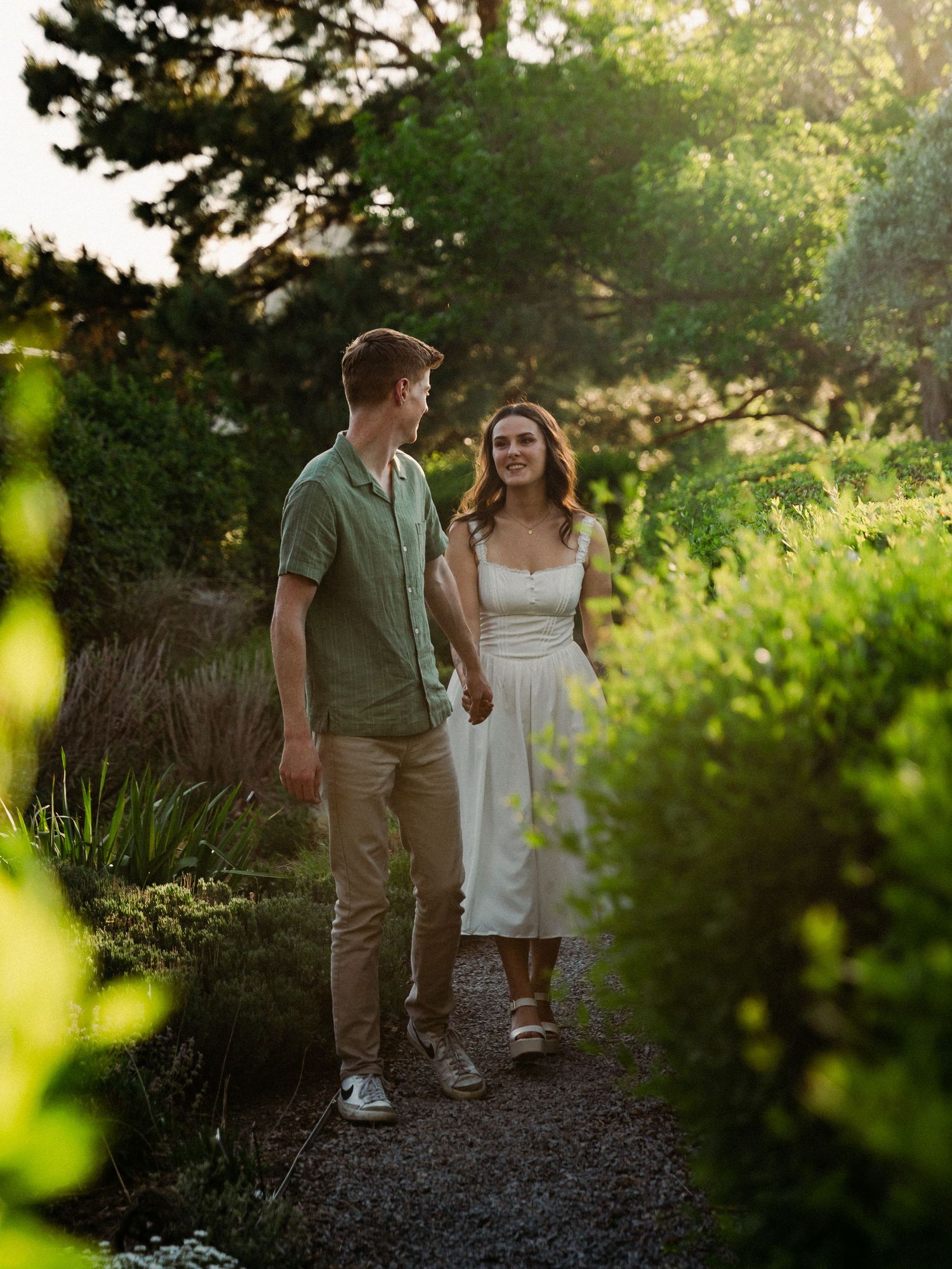 Couple walking hand in hand through a lush garden path at golden hour, man in green shirt, woman in white dress.