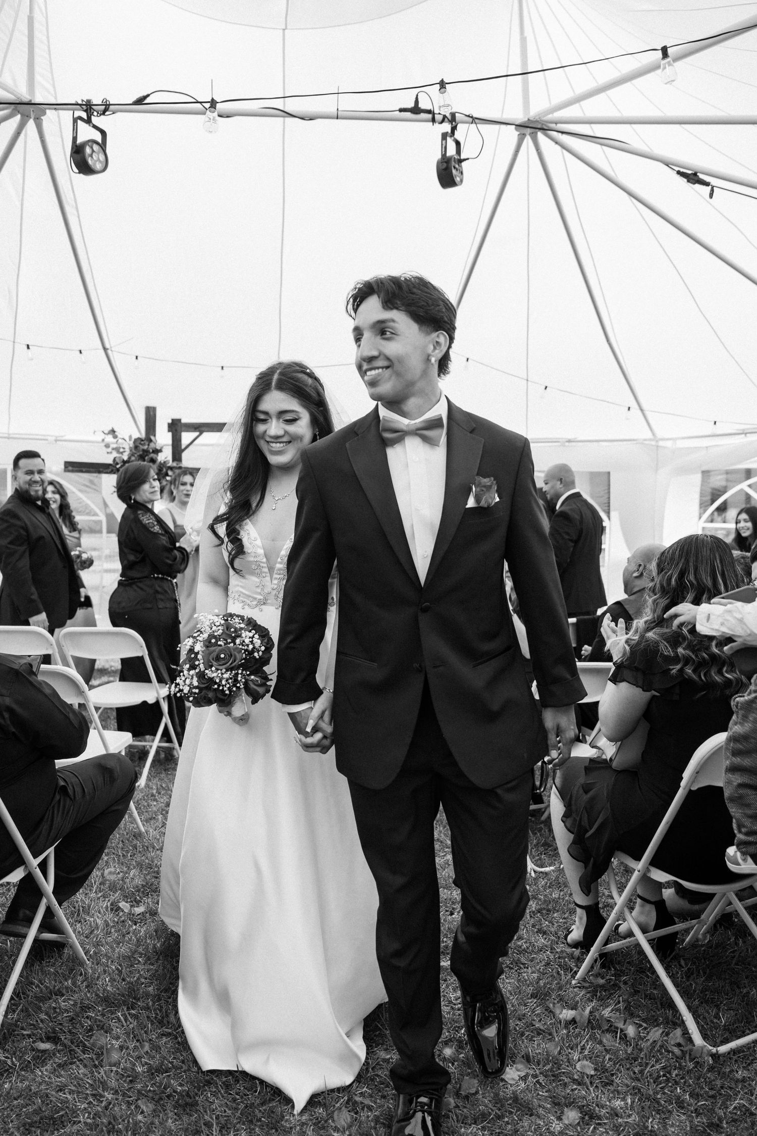 Black and white photo of a smiling bride and groom walking down the aisle under a tent at their wedding.