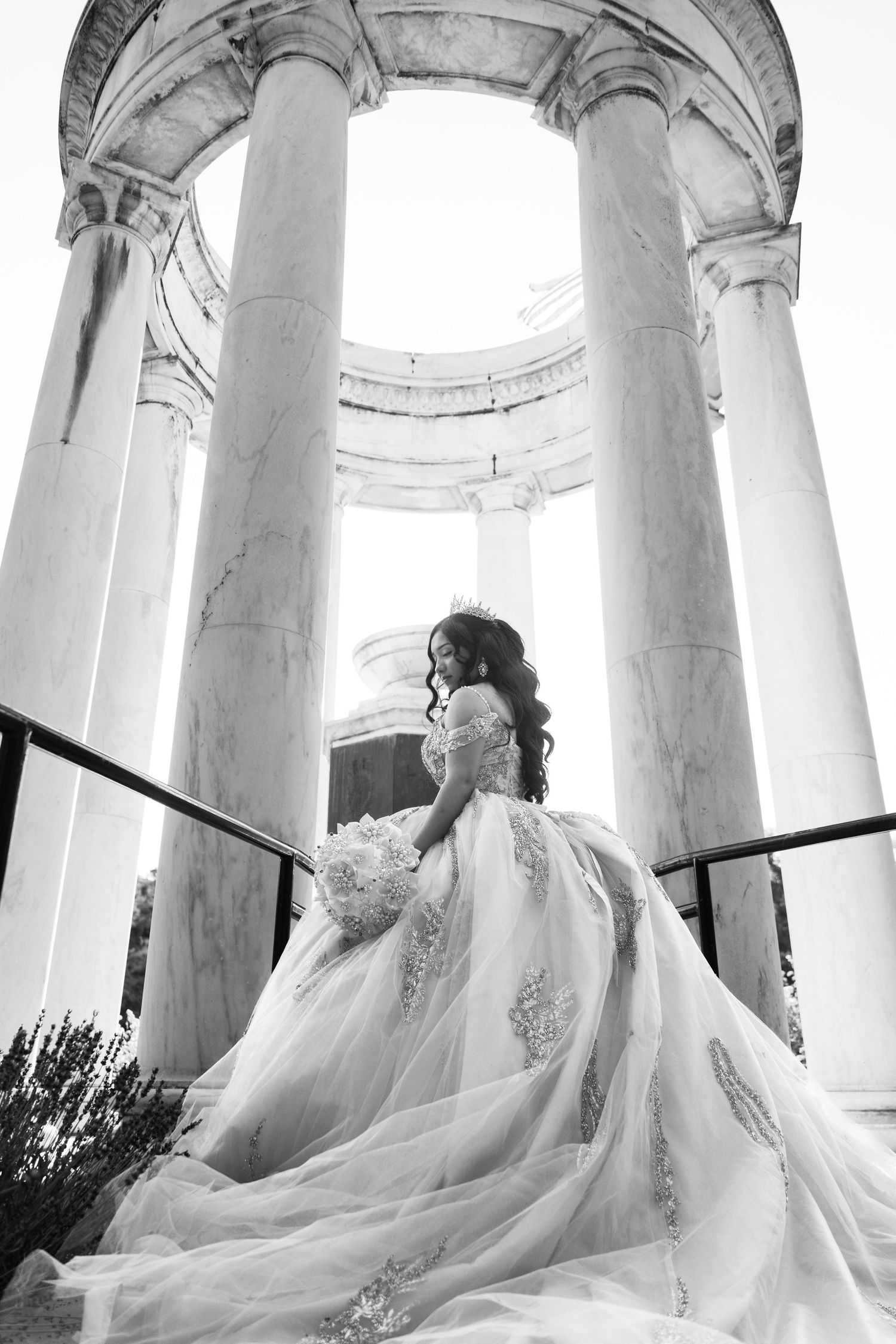 Black and white photo of a bride in a flowing lace gown holding a bouquet among grand marble columns.