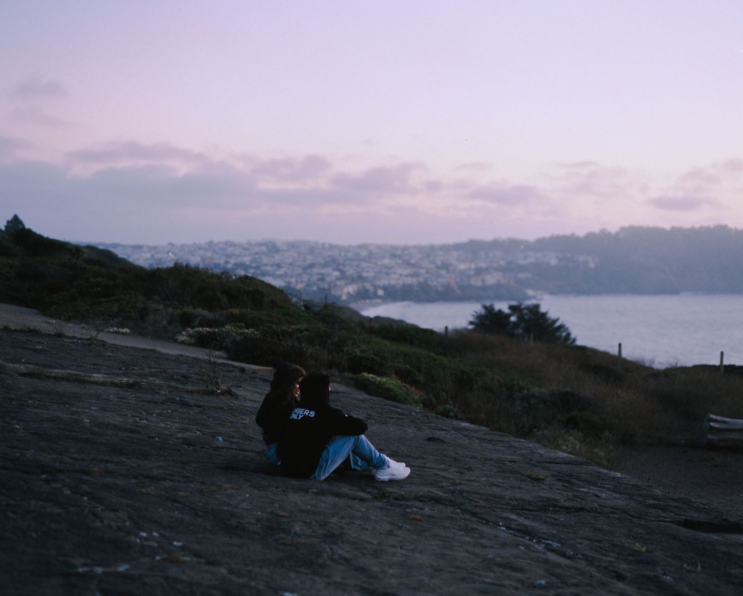 A couple sits together on a rocky hillside overlooking a coastal city at twilight.