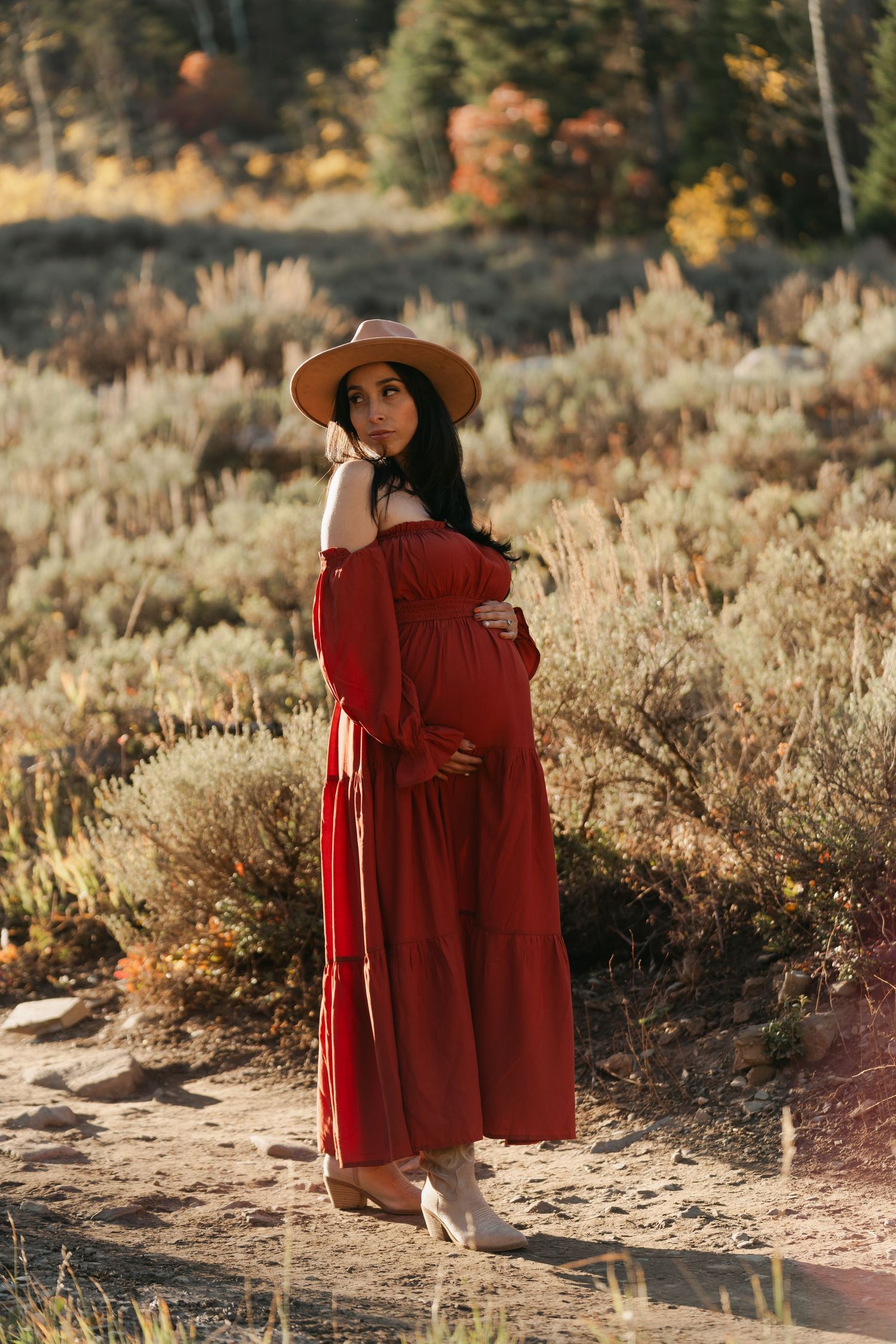 Pregnant woman in red off-shoulder dress and hat posing in autumn desert landscape.