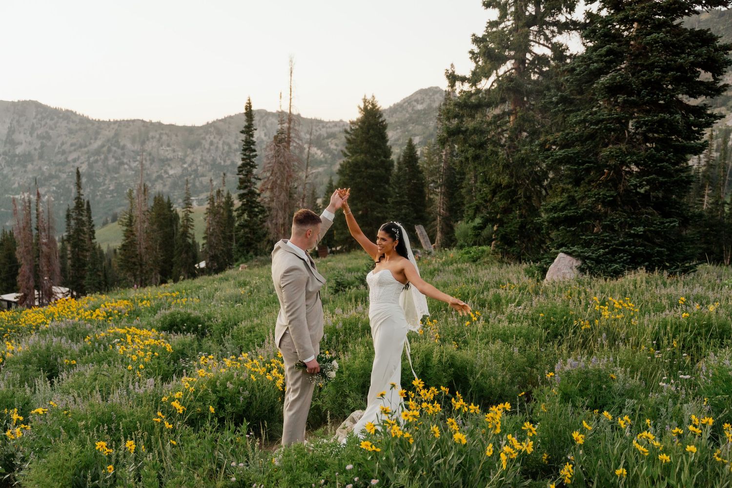 Bride and groom celebrating in wildflower meadow with mountain pine forest backdrop at golden hour.