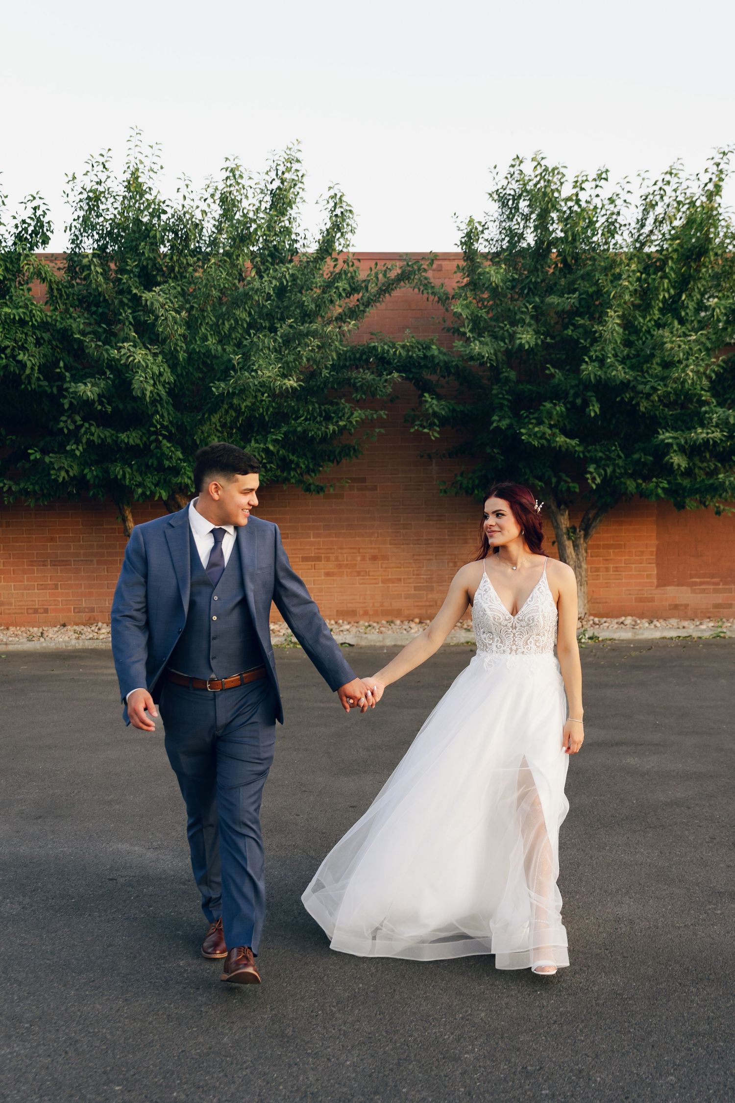 A bride and groom hold hands while walking outdoors in front of a brick wall with lush green trees.