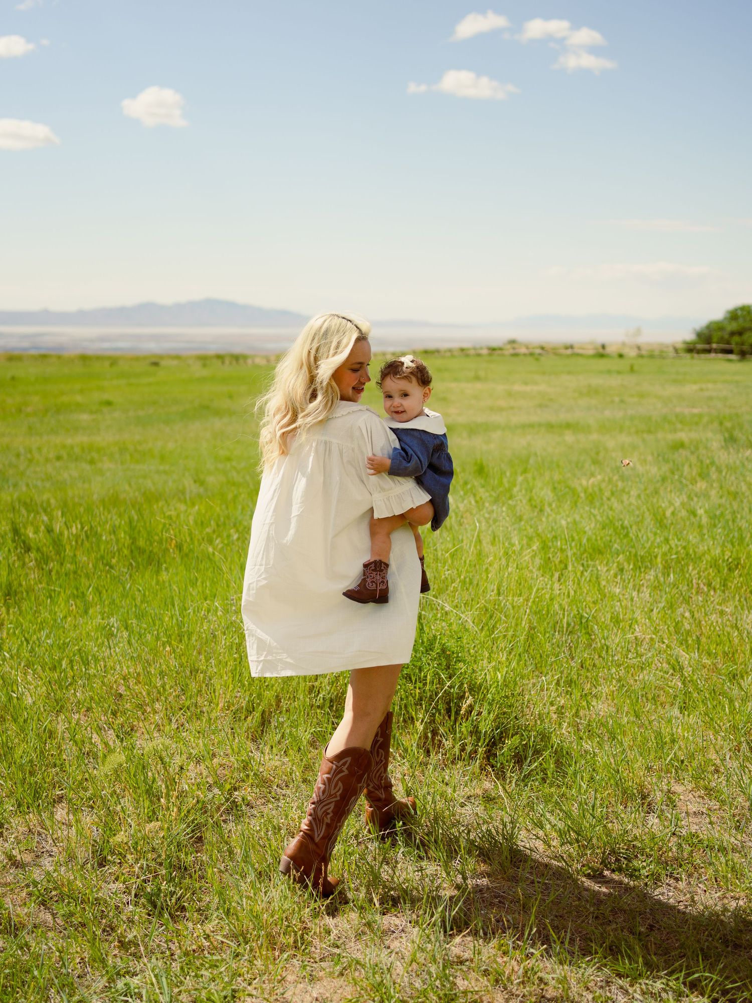 A blonde woman in a white dress and brown cowboy boots holds a baby in a green meadow under a blue sky.