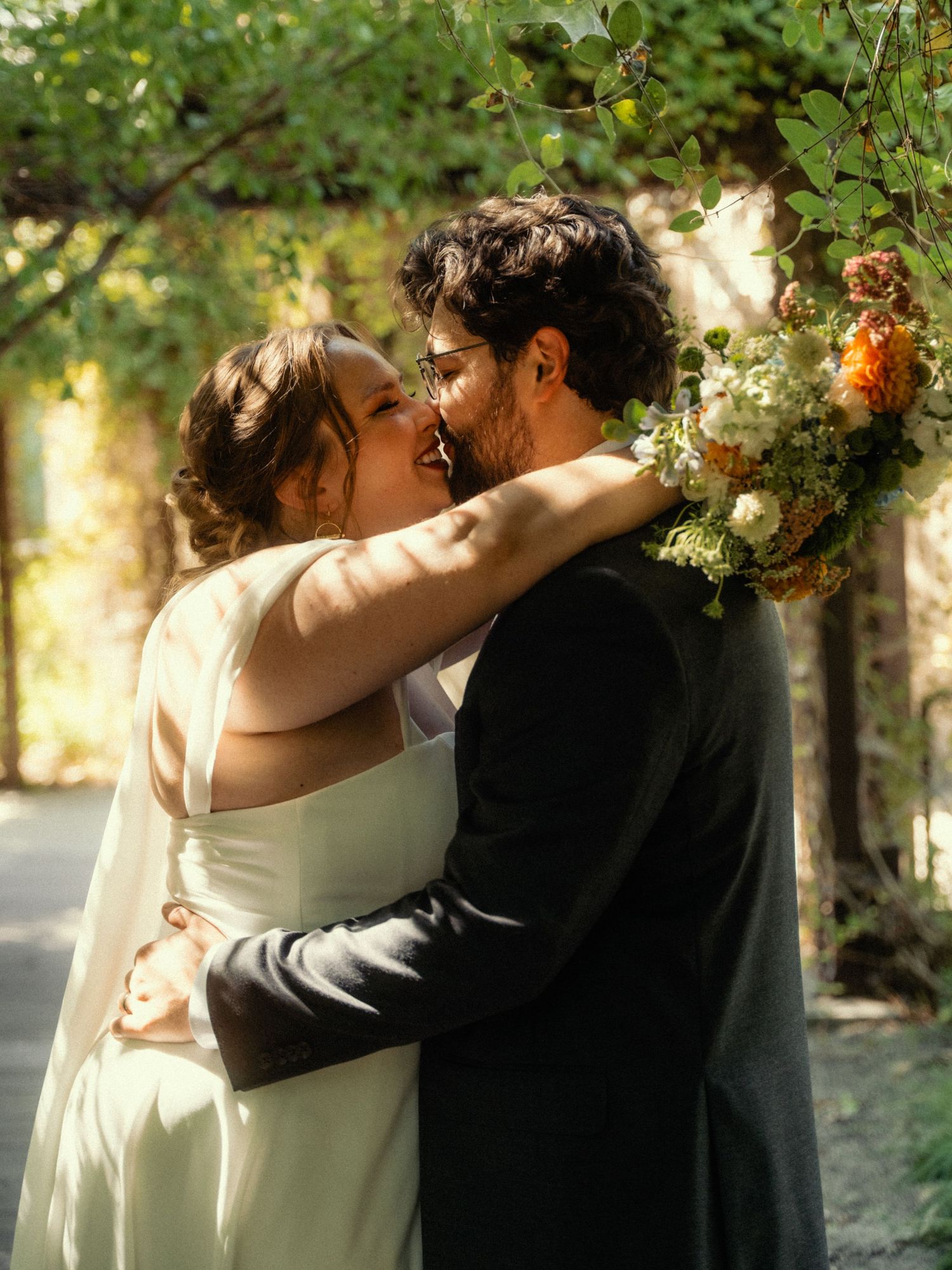 A bride and groom share a romantic kiss outdoors, surrounded by lush greenery and a beautiful floral bouquet.