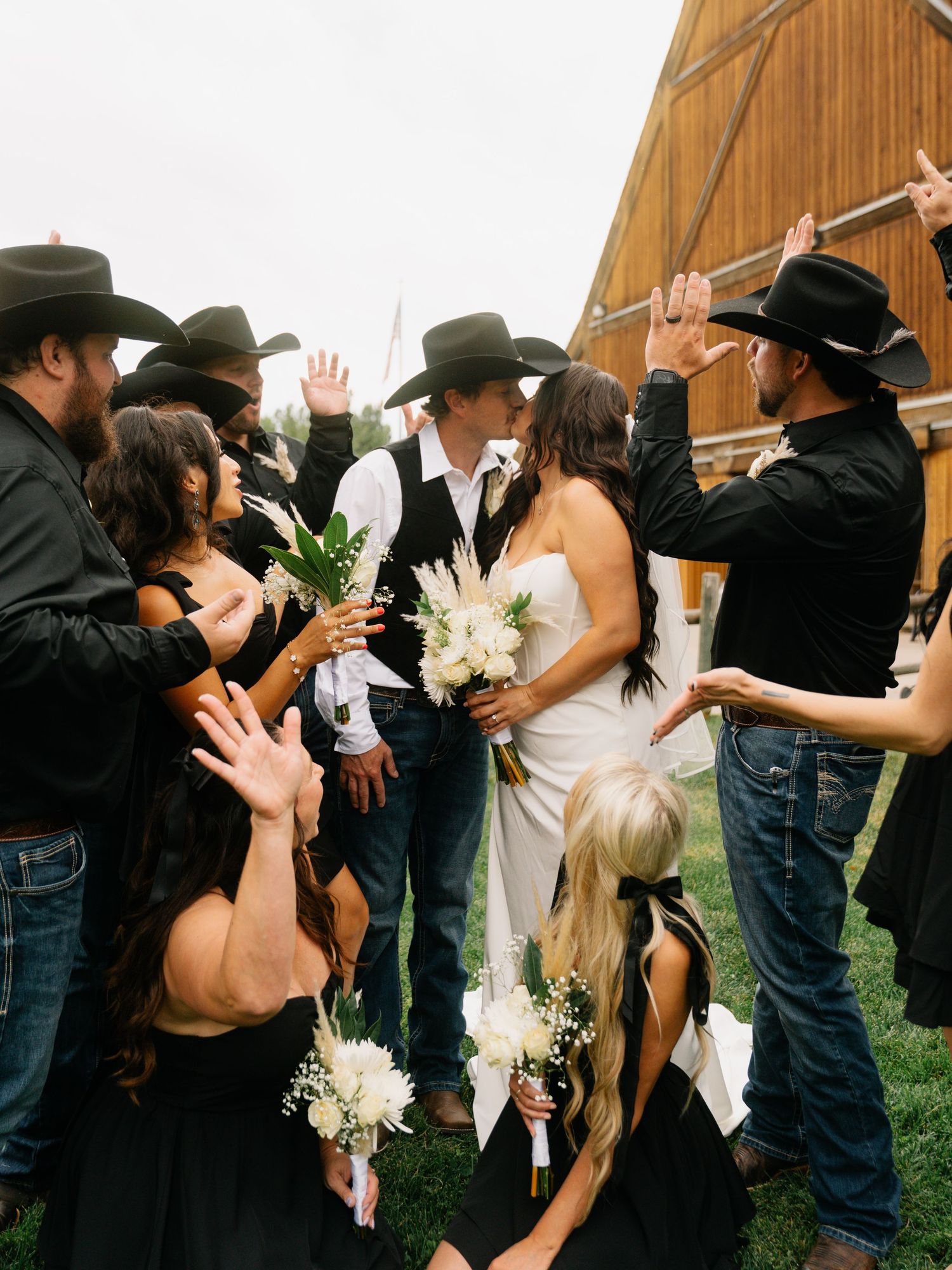 Country wedding couple kissing surrounded by guests in black cowboy hats holding white bouquets near a barn.