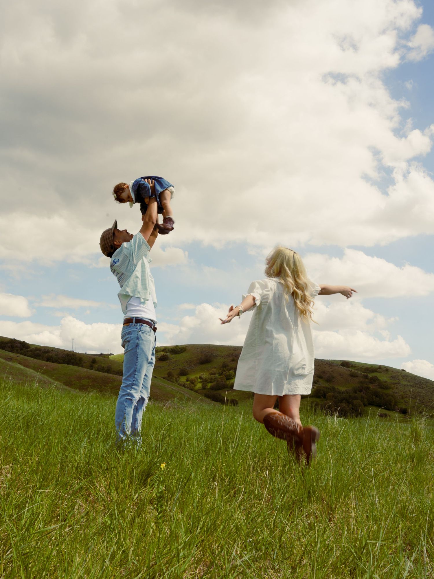 Family playing outdoors in green meadow, father lifting baby while girl in white dress runs joyfully under cloudy sky.