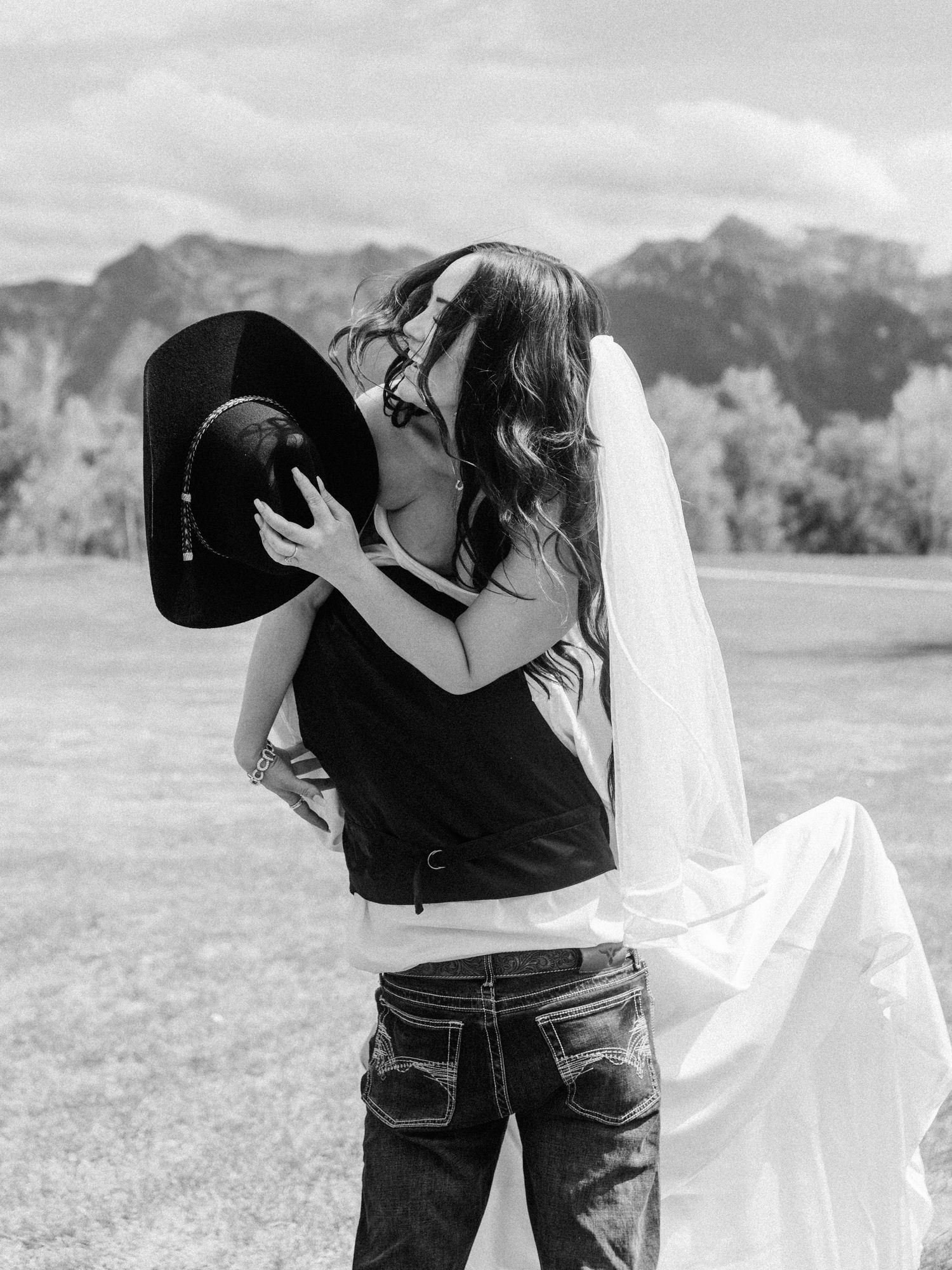Black and white photo of bride and groom kissing, cowboy hat, wedding veil, mountain backdrop.
