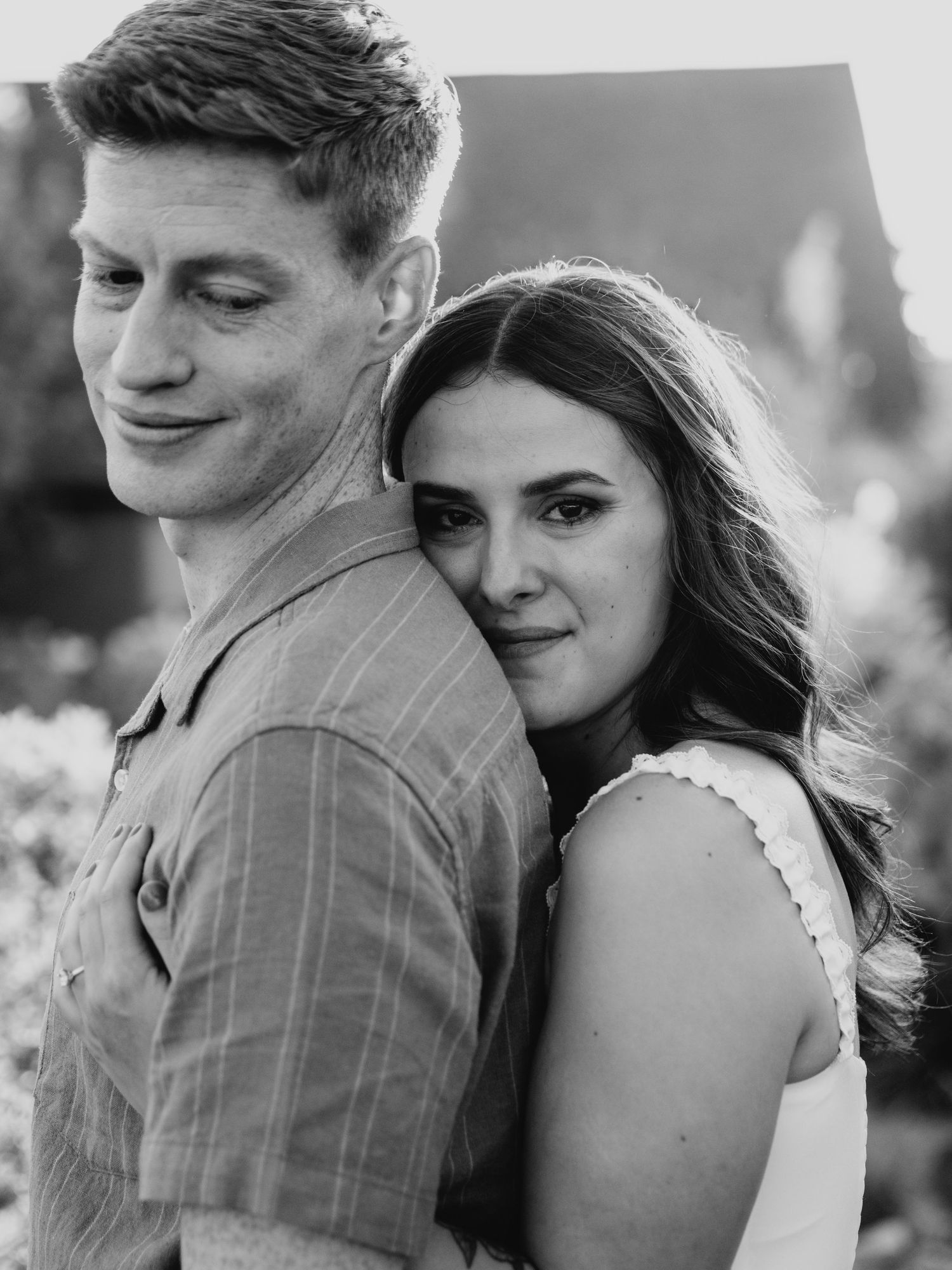 Black and white photo of a couple posing together, woman resting her head on mans back outdoors.