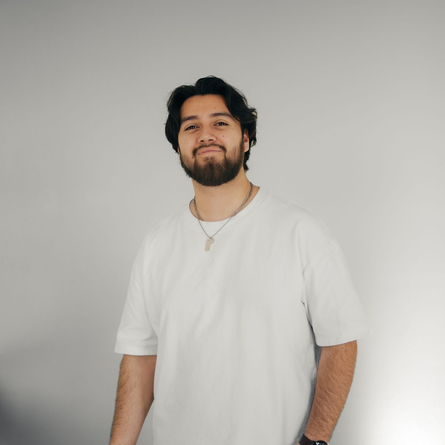 Young Hispanic man with beard wearing a white t-shirt posing against a light gray background.