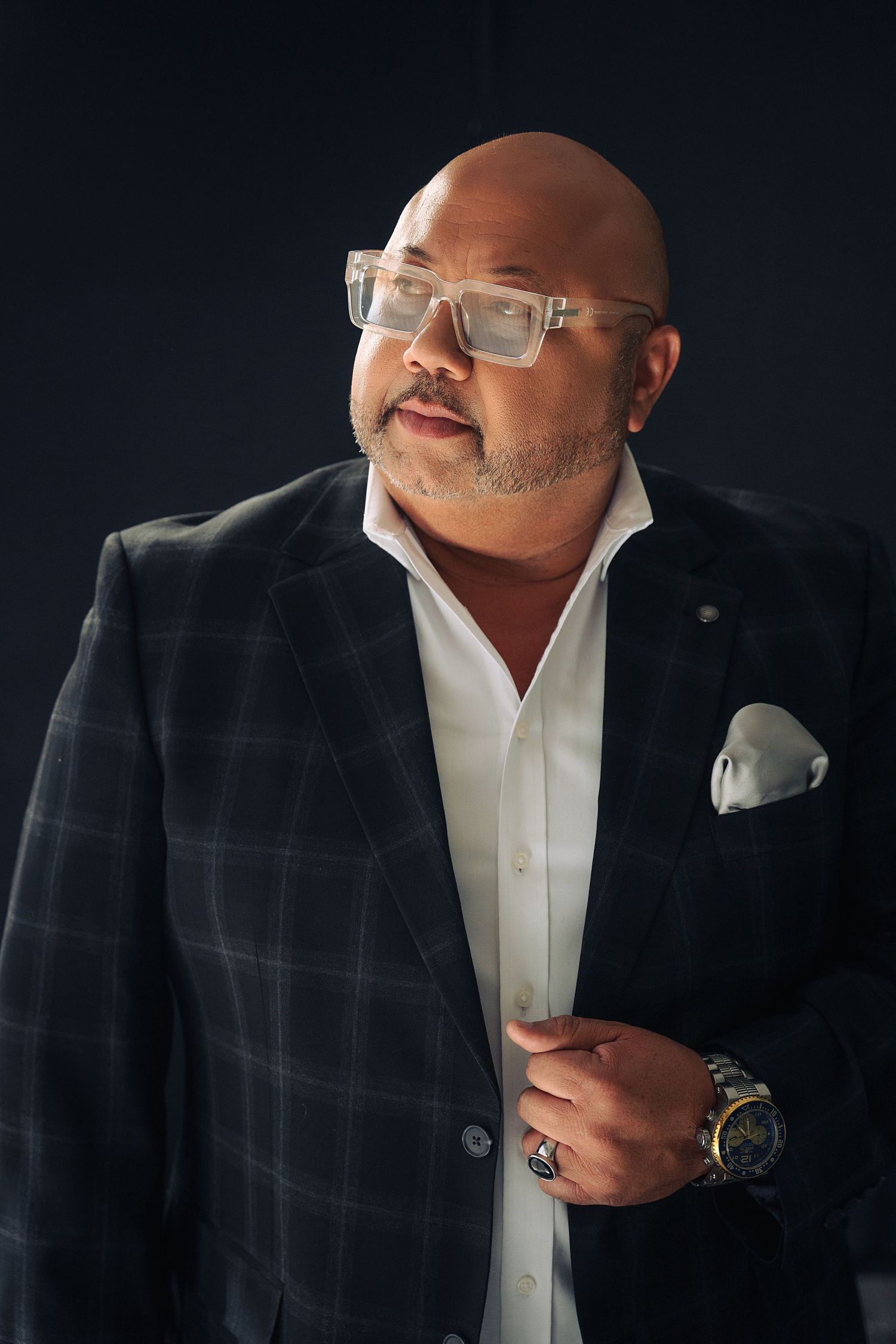 Bald man in plaid blazer with glasses and luxury watch posing confidently against dark background.