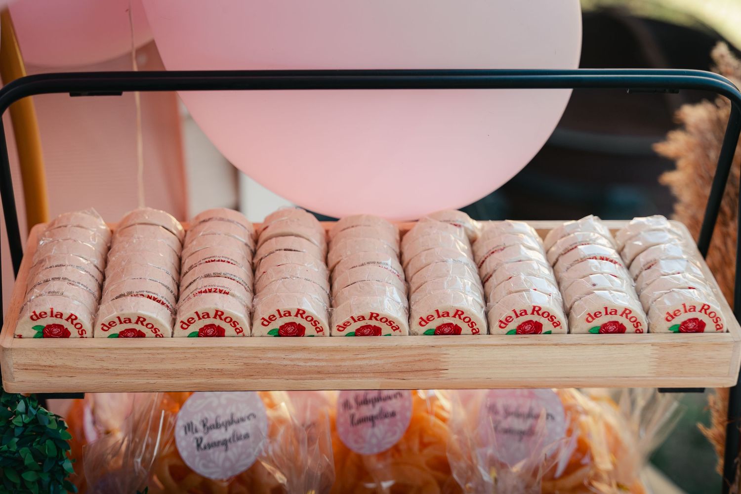 Rows of macarons with branded labels displayed on a wooden rack with pink balloons at a celebration event.