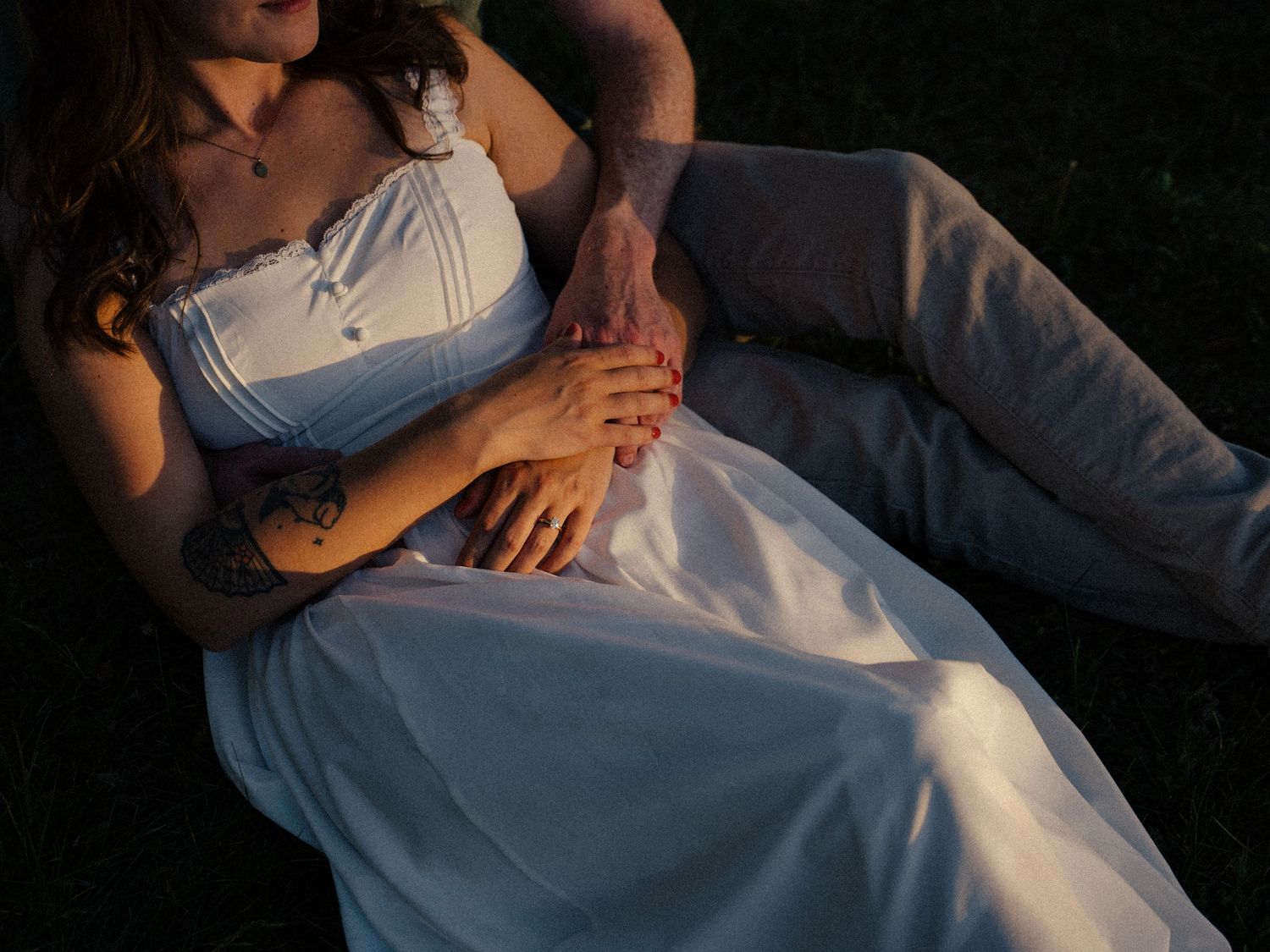 Couple in wedding attire lying on grass, bride in white gown with tattoo, groom in gray suit holding hands.