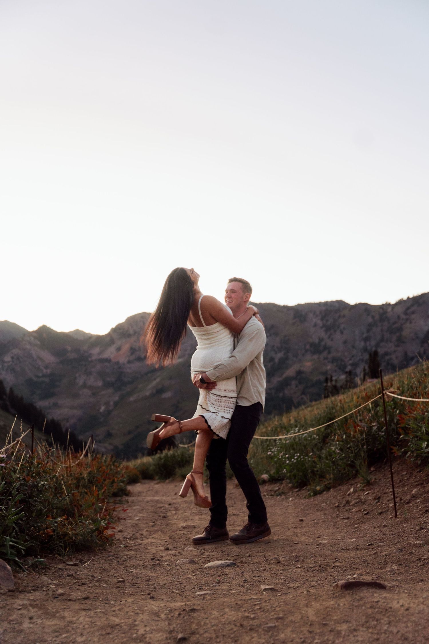 A couple embraces on a mountain trail at dusk, with scenic rocky peaks visible in the background.