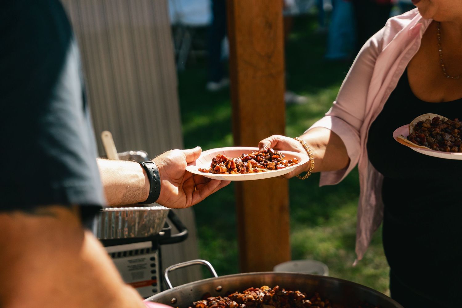 Person serving a plate of food at an outdoor catered event with a large pan of food.
