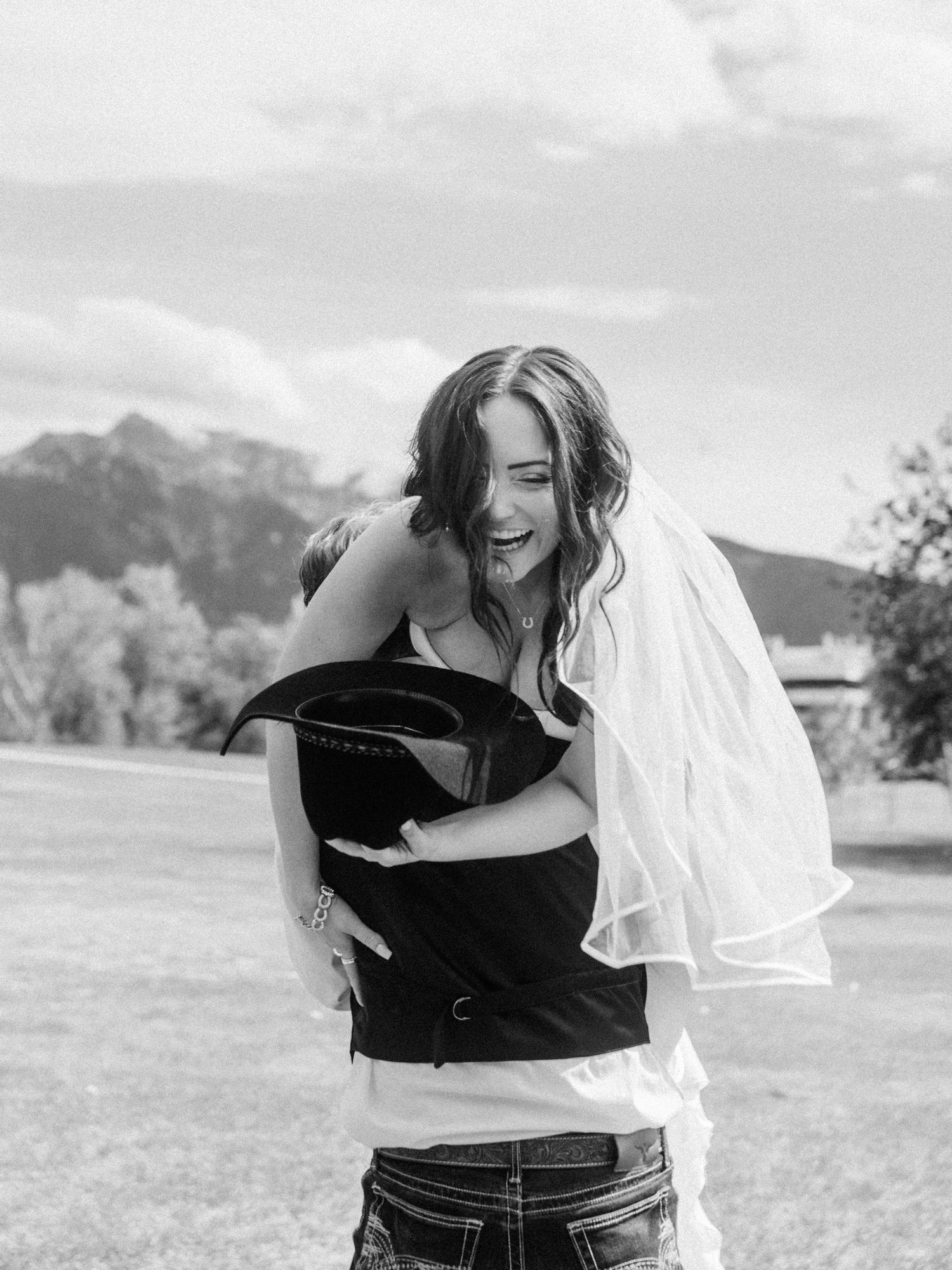 Black and white photo of a laughing bride in a veil holding a cowboy hat outdoors with mountains in the background.