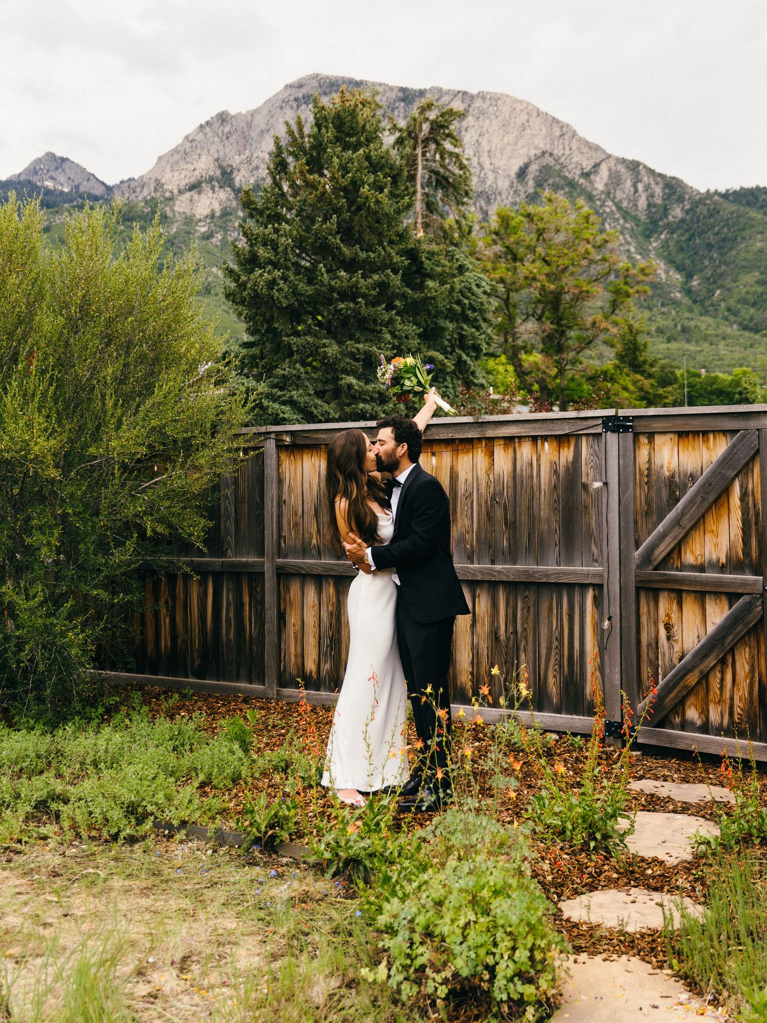 Couple sharing a romantic kiss in wedding attire with a wooden fence and mountain backdrop in a lush garden setting.